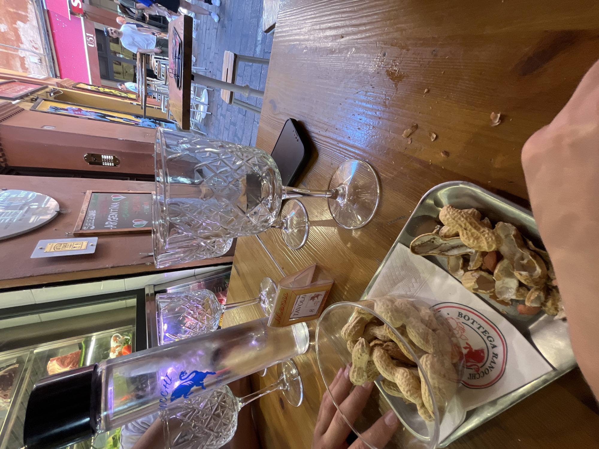 The image shows a table at a restaurant or bar in Bologna, Italy. The scene is captured from a high-angle perspective, looking down at the tabletop. Present on the table are several clear, ornate wine glasses, some empty and others possibly used. A small, rectangular black smartphone rests on the wooden surface of the table. A small paper menu card sits amongst the glasses. A silver tray contains a collection of peanuts, some still in their shells and others already opened. A clear plastic container also holds peanuts. A partially visible hand is present in the foreground of the scene. The environment suggests an outdoor seating area in the city, with pedestrians walking by and storefronts visible in the background.