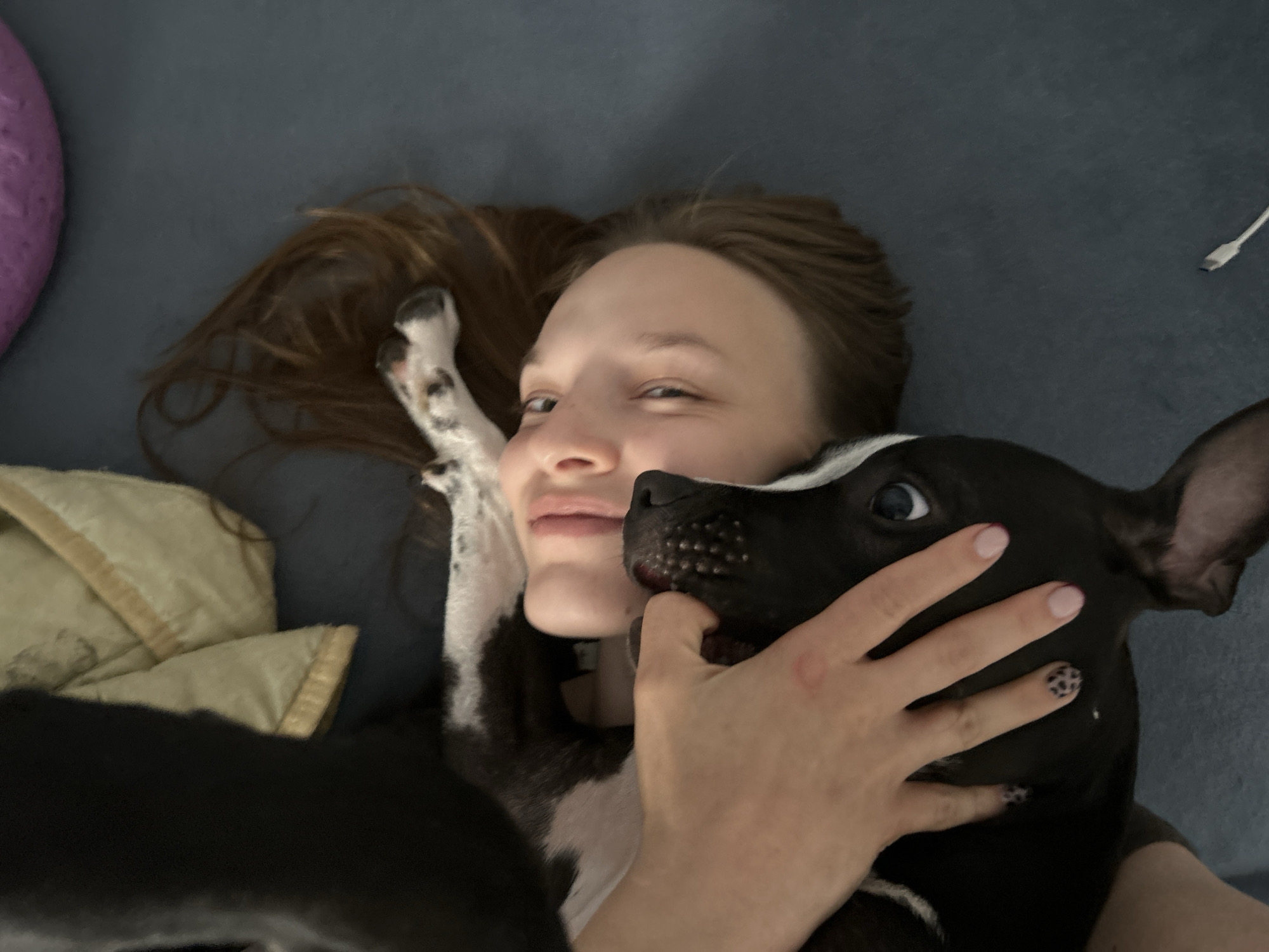 A young woman is lying on a dark blue surface, possibly a couch or bed, with a small black and white dog. She has her hair spread out behind her head, and she is looking at the camera with a playful expression, one eye winking. The dog is nestled close to her face, with its nose touching her cheek. The woman's hand is gently resting on the dog's face, and her fingernails are painted with a leopard print design. A blanket with a tan and cream pattern is visible to the left of the woman. In the background, on the right side of the image, a white USB cable is visible. The overall impression is one of intimacy and affection between the woman and her pet. The location in the city of Chaikovsky, Russia is not discernible from the image.