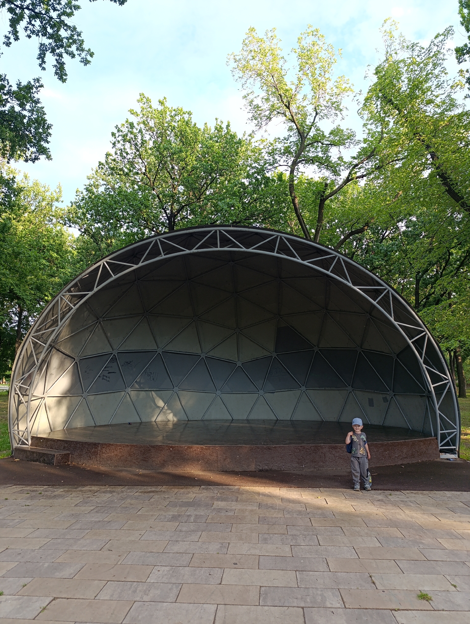 A young boy stands on the paving stones in front of a semi-domed outdoor stage in Novokuybyshevsk, Russia. The stage has a weathered wooden platform and a geodesic dome-like structure behind it. The boy is wearing a grey t-shirt with a graphic print, grey pants, a blue baseball cap, and sneakers. He is looking at the camera and smiling, with a blue backpack slung over his right shoulder. Sunlight casts shadows on the stage and the paving stones. The background is filled with lush green trees, indicating a park or recreational area. The sky above is a light blue with scattered clouds.