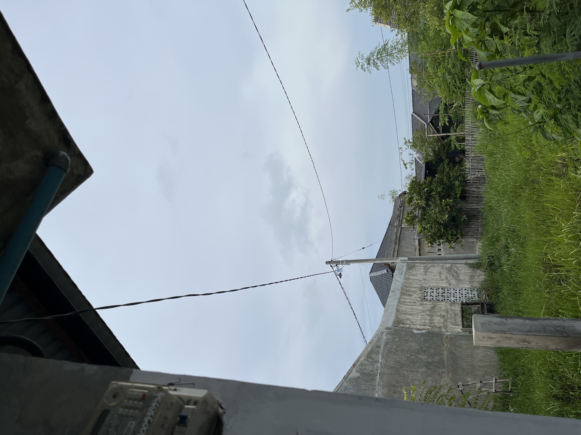 The image depicts a residential area in Lhokseumawe, Indonesia, under an overcast sky. In the foreground, a concrete pillar stands amidst overgrown green grass. To the left, a portion of a grey concrete building is visible, with an electrical meter attached to its wall. A wooden ladder leans against this structure. Further back, a fence made of bamboo palings separates the immediate area from neighboring houses. These houses have grey tiled roofs and concrete walls, with some showing patterned window openings. Several trees and dense vegetation are scattered throughout the scene. Electrical wires are strung across the sky, supported by a utility pole visible on the right. The overall impression is one of a developing or somewhat unkempt suburban environment.