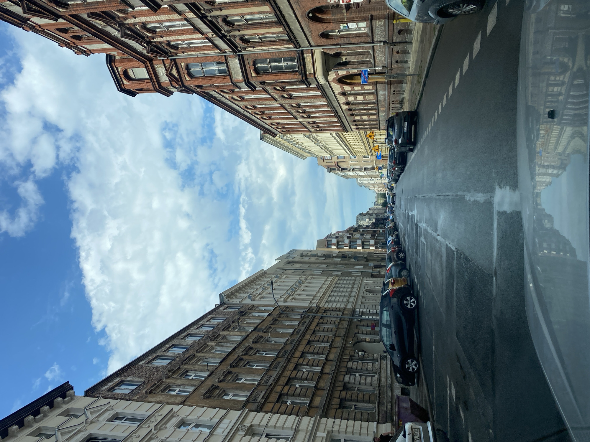 The image depicts a street scene in the city of Szczecin, Poland, likely in an older, residential area. The perspective is from inside a vehicle, looking forward down a wet asphalt road. On either side of the street are multi-story buildings, characteristic of European architecture, with ornate facades, arched windows, and brickwork. The buildings on the left appear older and more weathered, while those on the right seem to be in better condition.
Parked cars line both sides of the street. Several vehicles are visible, including a black hatchback, a yellow compact car, and other dark-colored sedans and SUVs. The wet road surface reflects the sky and the buildings, suggesting recent rain or damp conditions. The sky above is partly cloudy with a mix of blue sky and white clouds.
In the foreground, the dashboard and windshield of the vehicle are partially visible, reflecting the surrounding environment. A glimpse of a person can be seen on the left side of the street, near the parked cars. The street is relatively narrow, with a single lane of traffic visible in each direction, separated by white dashed lines. Streetlights are present along the sidewalks. The overall impression is of a typical urban streetscape in a European city on a cloudy day.
