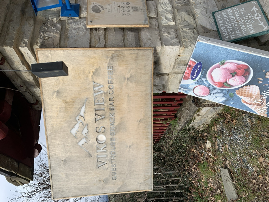 The image captures the exterior of a commercial establishment in Vikos, Greece, featuring multiple signs and traditional stone architecture. The main subject is a large, light-colored wooden sign prominently displaying "VIKOS VIEW" in a stylized font with a mountain logo above it. Below, the sign specifies "GUESTHOUSE SNACK BAR COFFEE."
To the right of this main sign, a colorful poster advertises ice cream from the "δωδώνη" (Dodoni) brand, showing scoops of pink ice cream with strawberries and waffle cones. Text on the poster includes "ΠΕΖΟ ΠΑΓΩΤΟ" (Pezzo Pagoto), suggesting a frozen dessert. Above the ice cream poster, a small green chalkboard sign lists available items: "ΑΝΑΨΥΚΤΙΚΑ" (Soft Drinks), "ΠΟΤΑ" (Drinks), "KAΦE" (Coffee), "(A)SYVEETS" (likely sweets), "ΚΑΣΙΟΠΙΤΑ" (Kasiopita, a type of pie), and "PIES."
Higher up on the stone wall, another small wooden sign is visible, bearing the word "DENTRO" and several pictograms, which could indicate services like Wi-Fi or pet-friendliness. The signs are set against a backdrop of a sturdy, light-colored stone wall, characteristic of traditional Greek architecture. A red metal railing is partially visible in the lower right, separating the immediate area from some sparse, dry vegetation on the ground. A black rectangular object, possibly a light or sensor, is attached to a wire above the "Vikos View" sign. The scene depicts an outdoor setting during daytime, inviting visitors to a guesthouse and cafe.