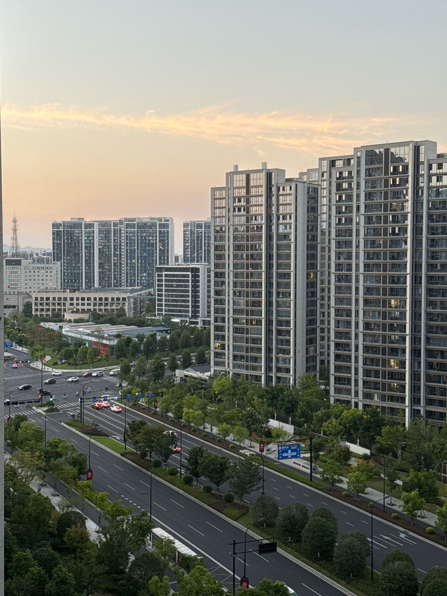 An aerial, eye-level shot captures a modern urban cityscape at dusk, characterized by tall, glass-fronted apartment buildings and a wide, multi-lane road. The sky is a soft gradient of pale yellow and blue, with a wispy cloud formation stretching horizontally across the upper portion of the frame. The buildings, with their reflective windows, are clustered together and dominate the background. In the foreground, the asphalt road is well-maintained, with clear lane markings and streetlights. Several cars are visible on the road, some with headlights on, suggesting the early stages of evening. Green trees and manicured landscaping line the road and surround the buildings, providing a contrast to the concrete and glass structures. Traffic signals and street signs are present at intersections. The overall impression is one of a well-developed, contemporary urban environment during twilight.