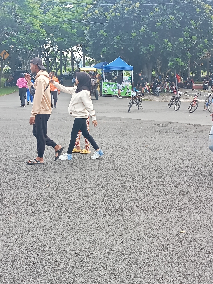 The image captures an outdoor scene in Singaparna, Indonesia, likely a public park or market area during the daytime with diffused light, suggesting an overcast sky or late morning/afternoon. The foreground features an asphalt road.
Two individuals are prominently walking from left to right. On the left, a man wears a beige hoodie, black pants, and sandals. His head is turned slightly to the left, and he appears to be holding something to his mouth, possibly a vape pen or small item. To his right, a woman in a beige hoodie, black hijab, black leggings, and blue and white sneakers extends her right arm forward, possibly gesturing or holding a device (not clearly visible) as if taking a photo or video. Another person's legs, wearing yellow flip-flops, are visible behind her.
In the mid-ground and background, more people are visible, some walking, others congregating near market stalls. There are several bicycles parked on the side of the road. Two market tents are present: a blue tent on the right and a yellow and white one further left. The blue tent has visible Indonesian text, including "300 RIBU BUNGA" (300 Thousand Flowers) and "JUAL BUNGA" (Sell Flowers), suggesting a flower vendor. Below the blue tent, a green stall has text that partially reads "MANTAN" or "MANTAP". A yellow road sign with a left-pointing arrow is partially visible on the far left. Lush green trees line the background, indicating a natural setting. The overall atmosphere is casual and bustling with activity.