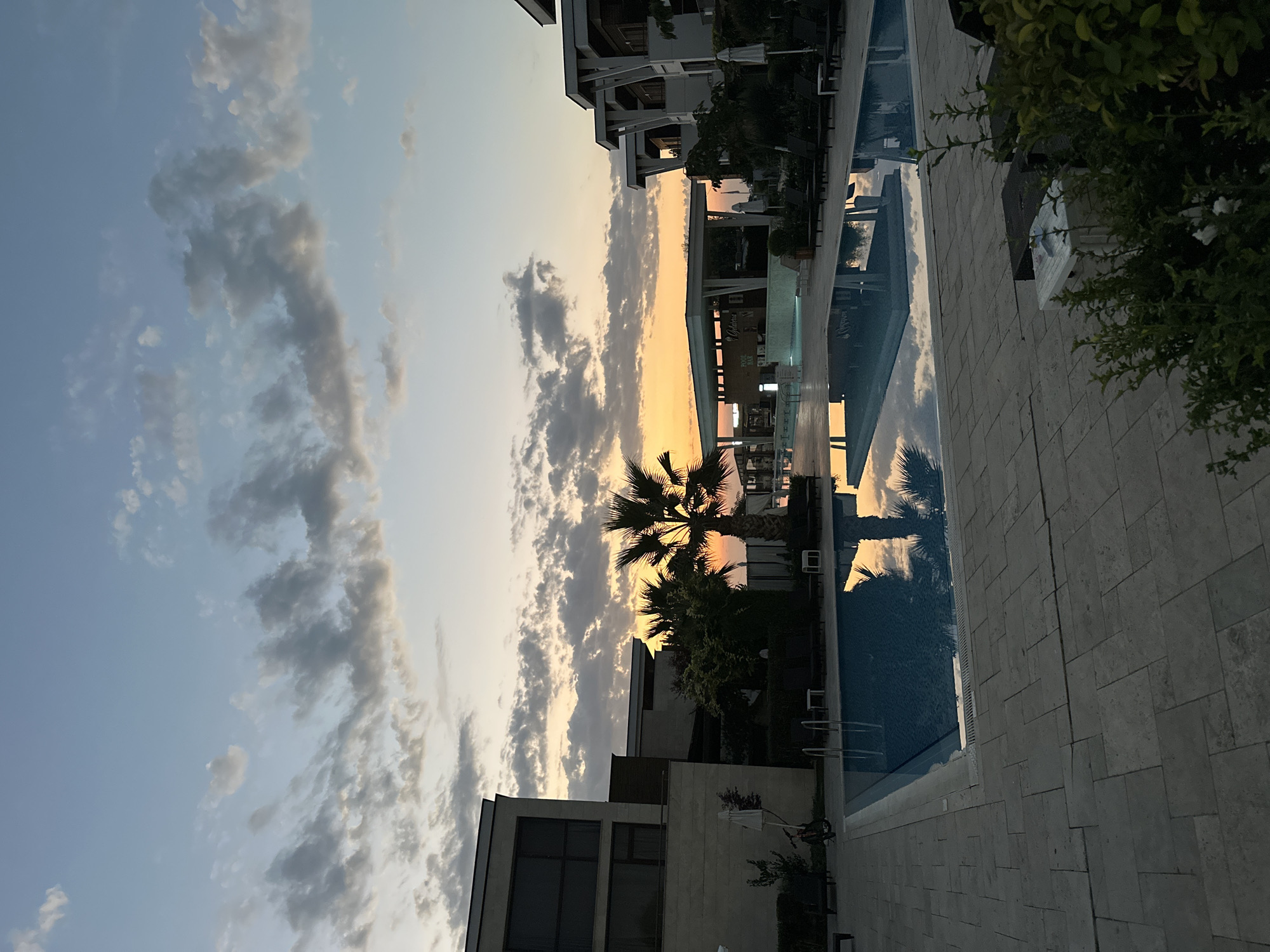 The image shows a resort with a swimming pool at sunset. The sky is a gradient of orange and blue, with scattered clouds. A palm tree stands in the foreground, silhouetted against the sunset. The resort buildings are modern and minimalist. The swimming pool is rectangular and empty, with lounge chairs surrounding it. The scene suggests a peaceful and relaxing atmosphere. The location is the city of Sozopol, Bulgaria.
