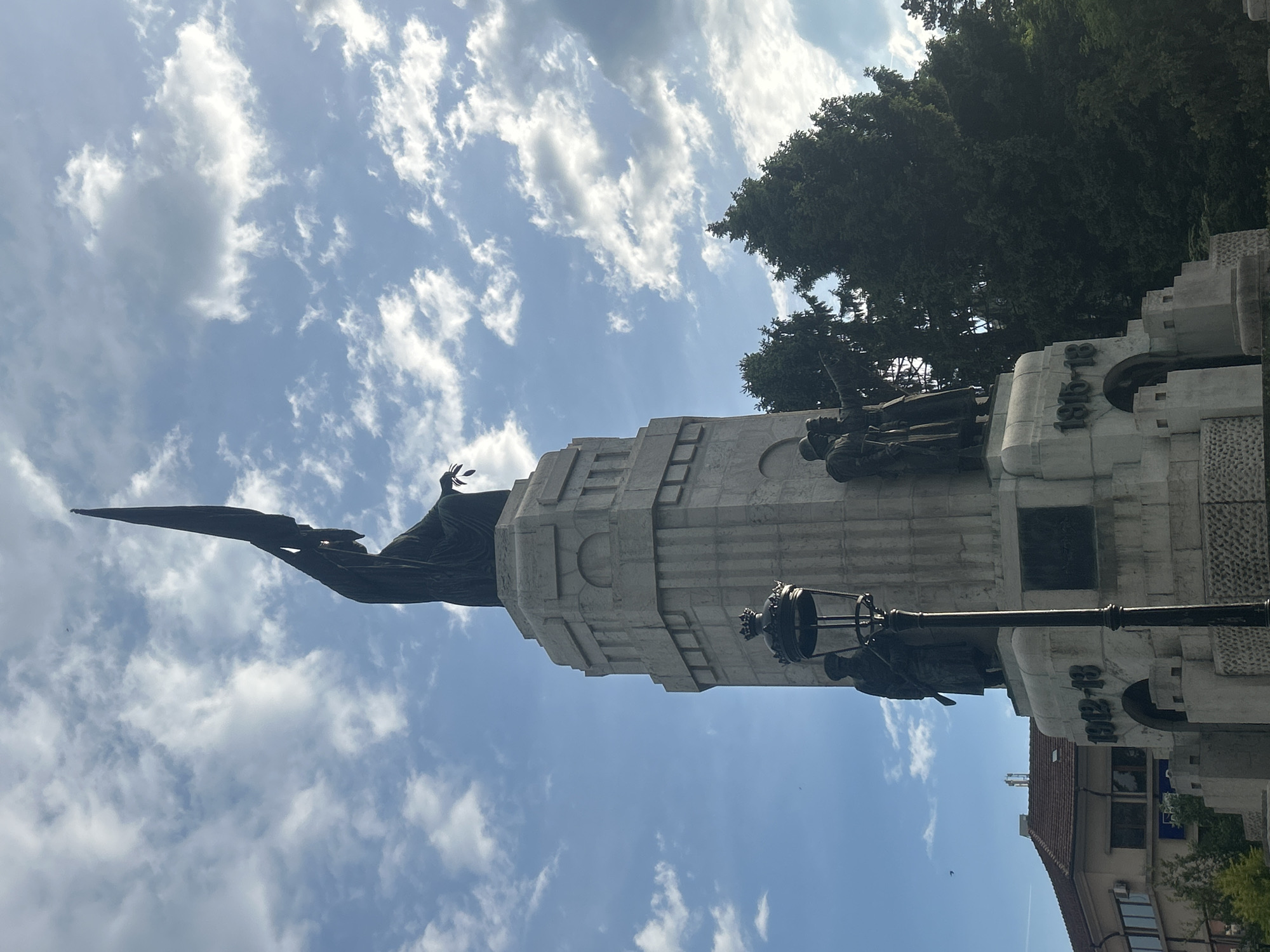 The image shows the Monument of Mother Bulgaria in Veliko Tarnovo, Bulgaria. The monument is made of white stone and features bronze statues of soldiers and other figures. The dates "1915-18" and "1941-45" are inscribed on the monument. The sky is blue with white clouds. A green tree is located behind the monument, and the roof of a building is visible in the foreground.