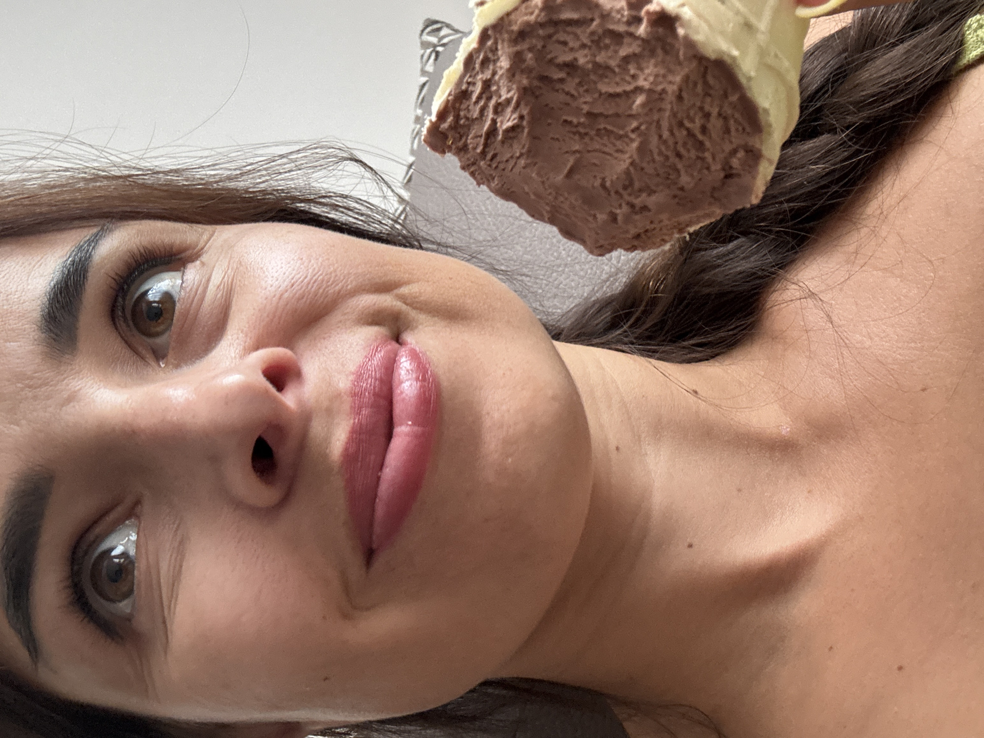 A close-up, medium-shot, waist-up photograph shows a woman looking up and to her left, with a piece of chocolate ice cream cone held near her mouth. The woman has brown eyes, dark brown hair, and rosy lips. Her skin is lightly tanned. She is wearing a green top. The background is a plain, light-colored wall. The lighting is natural and appears to be coming from the right. The woman's mouth is slightly open, and her lips are glossy. She has a slight smile on her face. The ice cream cone is partially eaten, with visible layers of chocolate ice cream and a light-colored cone. The image is sharp and in focus. The woman's body is angled slightly to the left. Her right arm is holding the ice cream cone, and her hand is visible in the lower right corner of the frame. The woman's left shoulder is visible, as well as her neck and upper chest. The ice cream is a rich, dark brown color. The cone is a light yellow or cream color. The ice cream has a textured surface, with visible swirls.