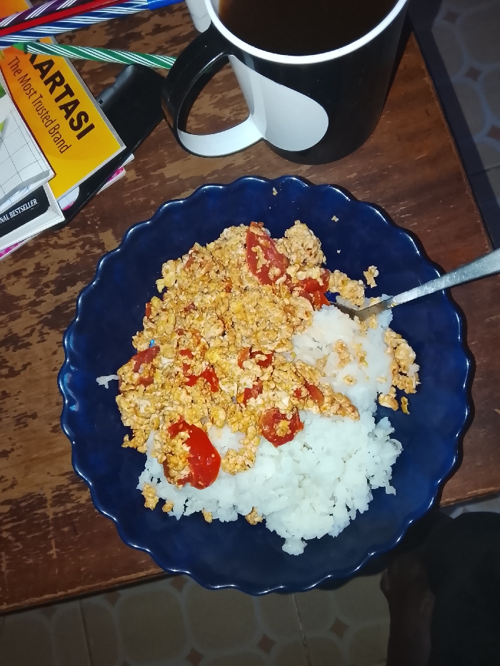 The image captures an overhead view of a meal and associated items on a wooden table within an indoor setting in Kiambu Township ward, Kenya. Dominating the center-right is a deep blue, fluted-edge ceramic plate containing white rice partially covered with scrambled eggs mixed with diced red tomatoes. A silver spoon is positioned in the food, scooping a portion of rice. To the upper-right, a black and white ceramic mug with a distinct white heart design holds a dark brown liquid. The upper-left quadrant features a stack of printed materials, with a yellow book prominently displayed, bearing the text "KARTASI The Most Trusted Brand." Scattered nearby are several multicolored pens (blue, red, green, purple, orange) and a dark electronic device. The floor, visible in the lower-right, consists of light-colored tiles arranged in a geometric pattern, alongside an indistinct dark object.