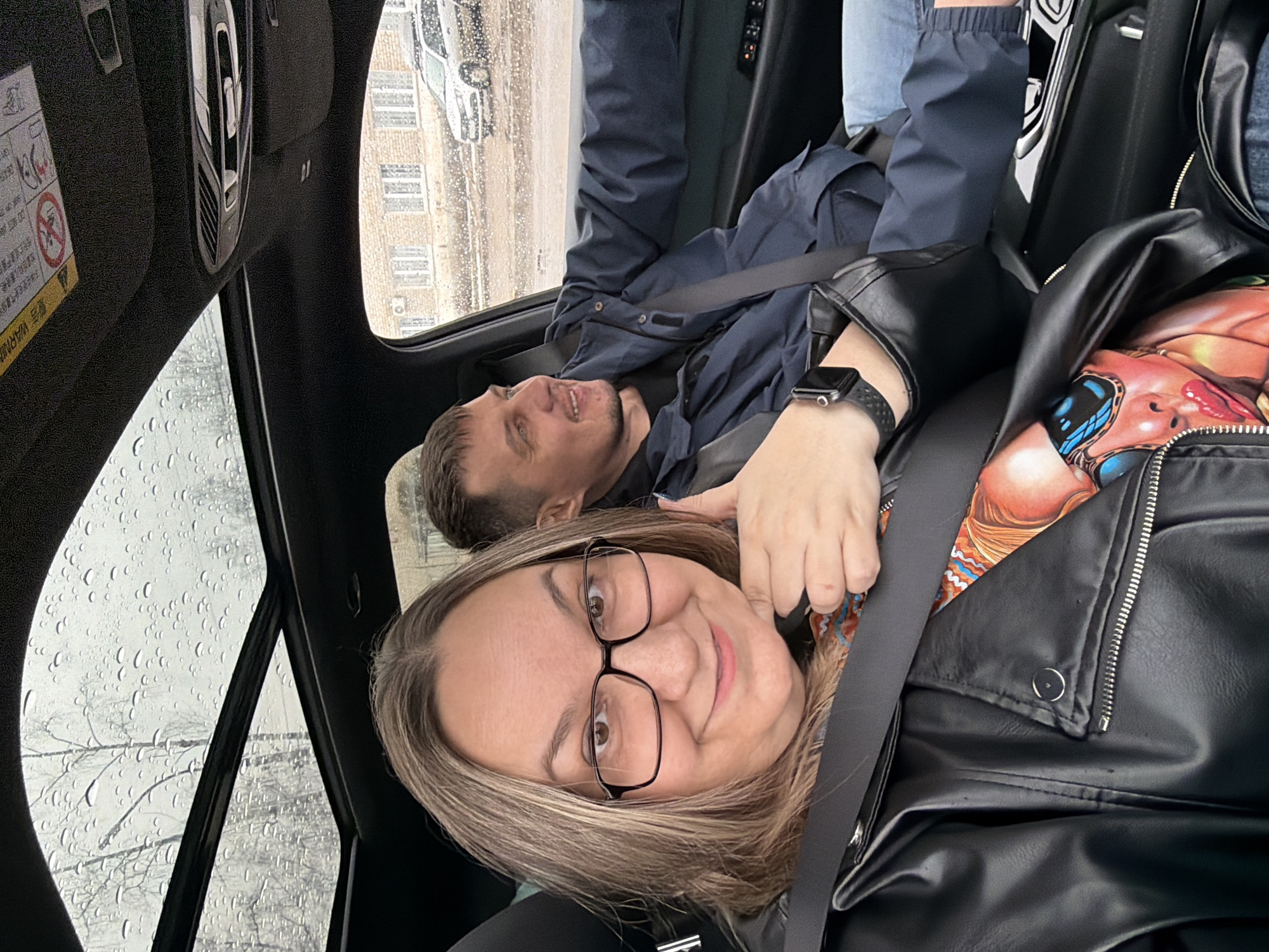 Two people are seated in a car. It appears to be raining outside. The woman in the foreground is wearing glasses and a black leather jacket. The man in the background is wearing a dark blue jacket. Both are wearing seatbelts. Based on the exterior view, the location appears to be in a city, possibly Chaikovsky, Russia. The view outside the window shows buildings. The windshield has rain droplets. The interior of the car is dark.