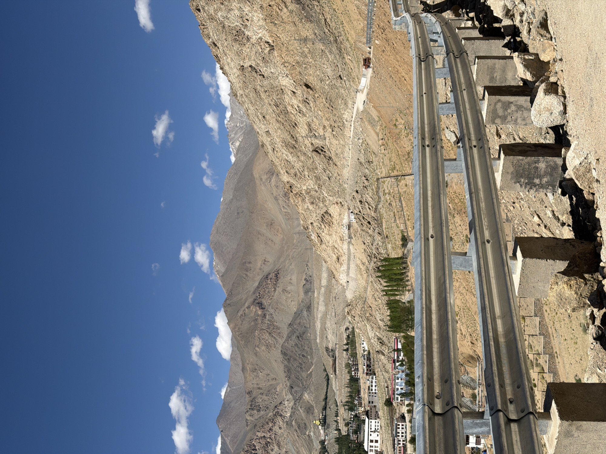 The image depicts a mountainous landscape near the city of Leh, India, likely on a road leading to a high altitude monastery or viewpoint. In the foreground, a silver metal guardrail supported by concrete posts lines a winding road. The road itself is paved with gravel and curves upwards into the rocky, barren mountainside on the right. In the mid-ground, a small village is nestled in a valley, characterized by white and beige buildings with red roofs, interspersed with clusters of green trees. A prominent golden statue of Buddha is visible on a hilltop overlooking the village, likely part of a monastery complex. Further back, massive, arid mountains dominate the horizon, their slopes etched with erosion patterns and sparsely vegetated. The sky is a vibrant, clear blue with a few scattered white clouds. Power lines and transmission towers are visible along the mountain slopes, indicating infrastructure in this remote area. A red truck is visible driving on the road in the distance. The overall impression is one of a remote, high-altitude environment with a developing settlement in a starkly beautiful natural setting.