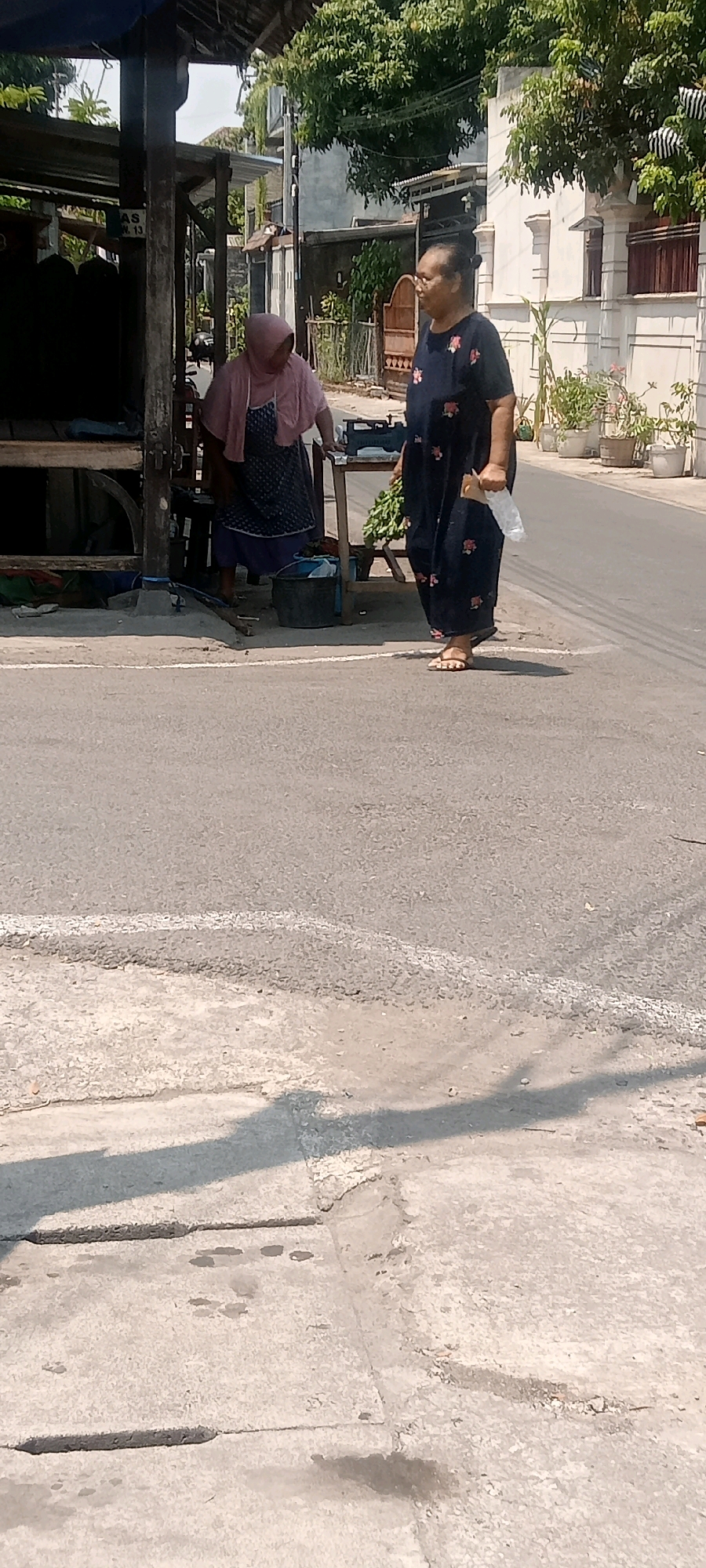 The image depicts a street scene in Surakarta, Indonesia, under bright daylight. Two women are present. One woman, wearing a pink headscarf and a patterned skirt, is standing behind a market stall. She is facing left, with her body bent slightly forward. A scale is visible on the stall in front of her. The second woman, wearing a dark blue patterned dress, stands on the right side of the frame, facing the first woman. She is holding a white plastic bag in her right hand and a bunch of green produce, possibly vegetables, in her left. The environment includes a street with a white line marking, a concrete drainage cover in the foreground, and buildings with trees and potted plants in the background. The overall atmosphere suggests a typical street market setting in Indonesia.
