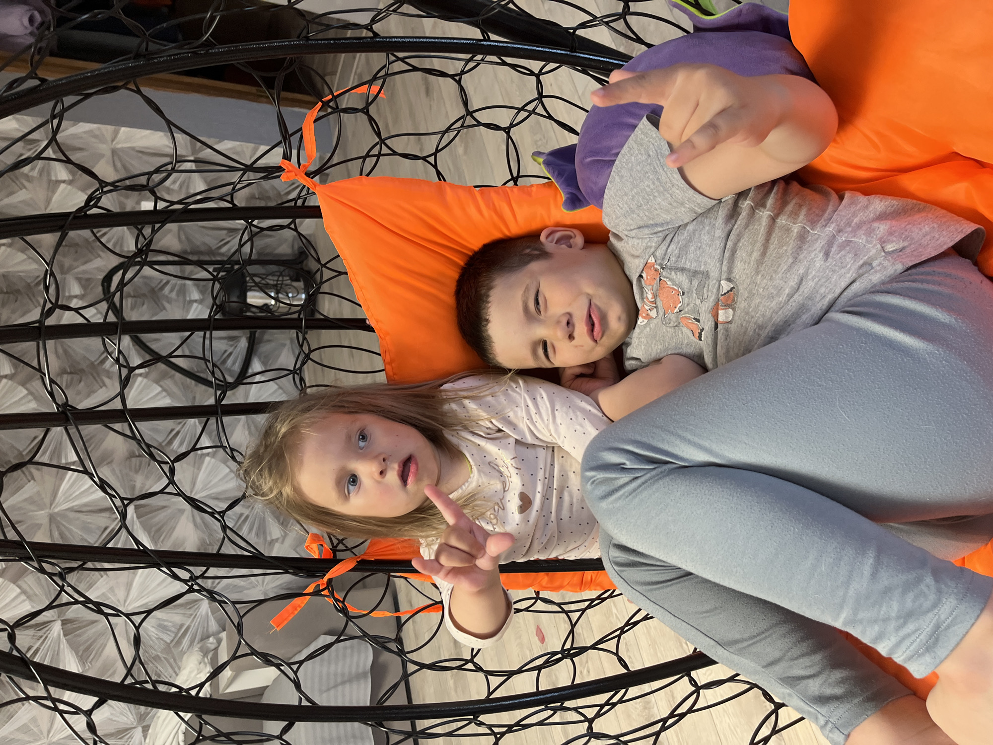 Two children are lying in a black metal hanging chair. The chair has an orange cushion and a purple pillow. A girl with blonde hair and a white shirt with brown dots is on the lower left, making a hand gesture. A boy with brown hair and a gray shirt with foxes is on the upper right, also making a hand gesture. The chair is positioned on a light-colored floor. The background is out of focus, but appears to be a wall with patterned wallpaper. The location is Chaikovsky, Russia.