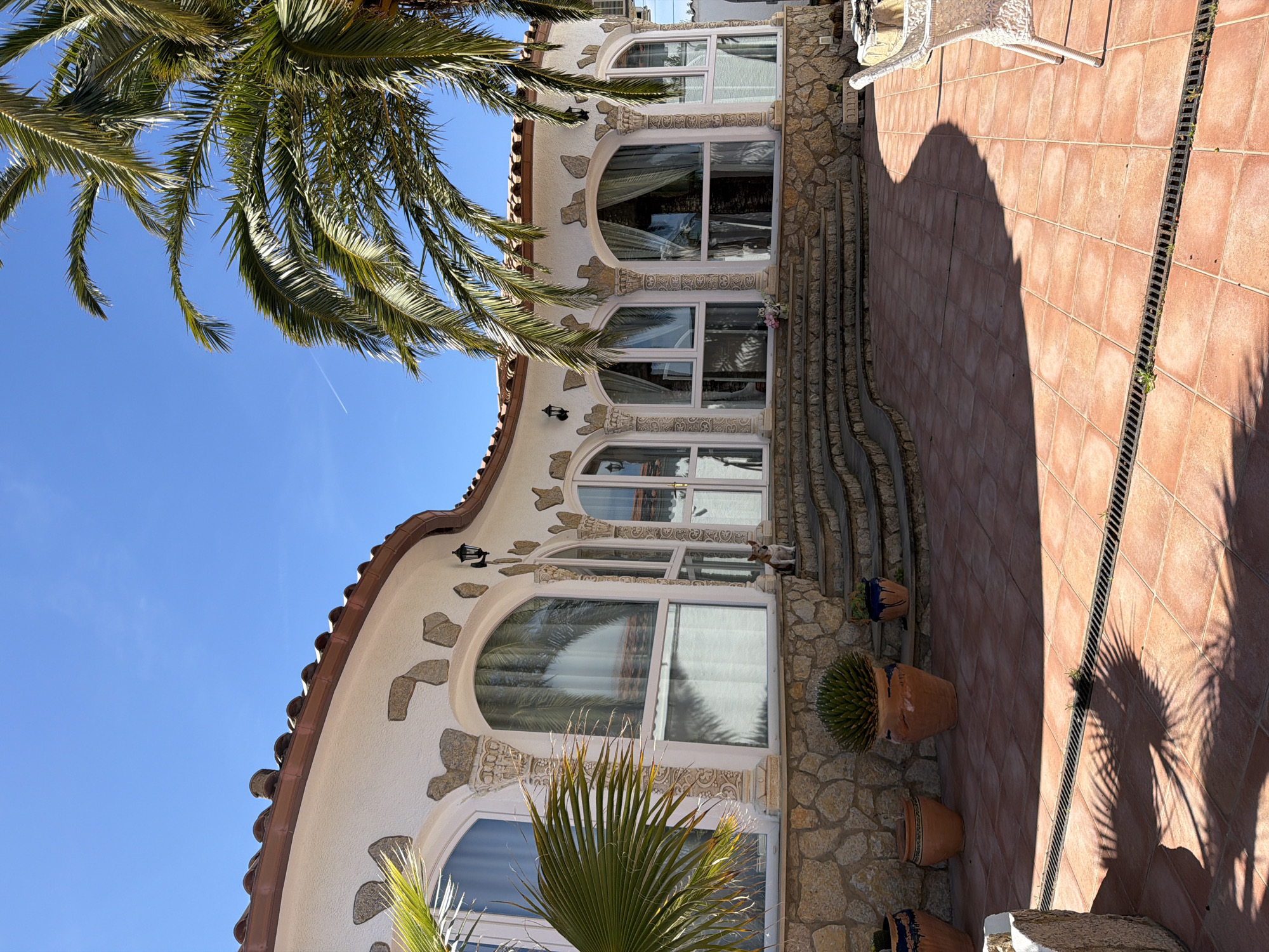 The image shows a curved building in Mont-roig del Camp, Spain, featuring a white facade with stone accents and arched windows. A tiled roof is visible, and the building appears to have multiple stories. Palm trees are in the foreground, partially obscuring the view of the sky and building. The ground is covered with red tiles and includes a drainage channel. Flower pots are placed along the building's base and stairs. A small dog statue is visible on the windowsill of one of the windows. A white wicker chair sits on the tiled area. The scene is brightly lit with strong shadows from the palm trees.