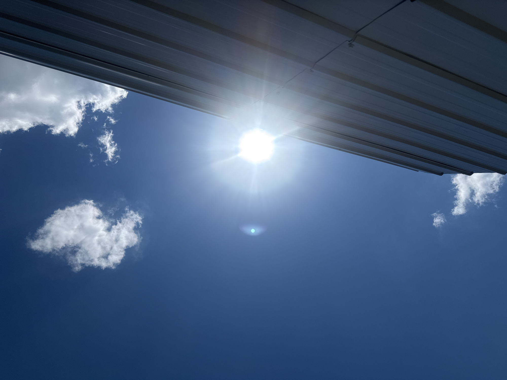 The image depicts a low-angle view of a bright sun shining against a clear blue sky. A portion of a modern building, possibly made of corrugated metal siding, occupies the right side of the frame, running diagonally from the lower right to the upper right corner. A lens flare from the sun is visible in the upper middle section of the sky. Several small, wispy white clouds are scattered throughout the sky, one prominent cloud cluster is near the upper left. The scene appears to be an outdoor environment within the Village of Johnson City, United States of America. There are no people visible in the image.