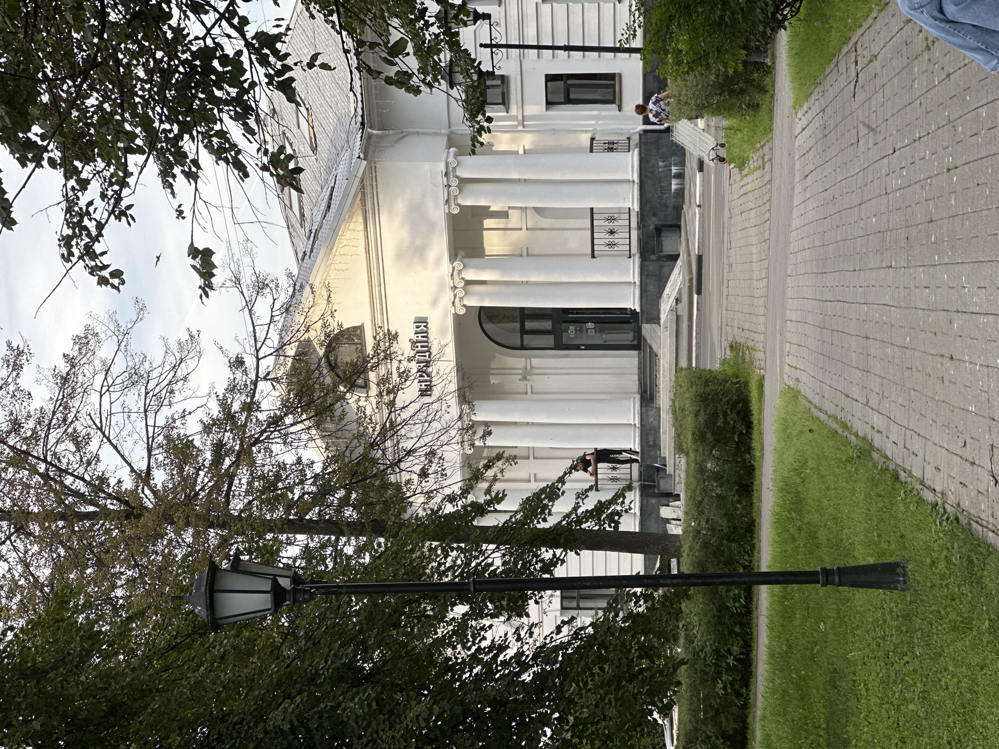 In Putyatino (Semivragovskaya S/A), Russia, a white building with a classical facade stands behind a paved walkway and manicured green bushes. The building features prominent white columns and an arched entrance. A black lamppost with an ornate design is positioned to the left of the walkway. Two people are visible: one individual stands near the building's entrance, leaning on a railing, and another person is seen pushing a wheeled cart on the right side of the walkway. Trees with green and yellowing leaves flank the scene, and the sky is visible above, with a bird in flight. The lighting suggests it is daytime, possibly late afternoon.
