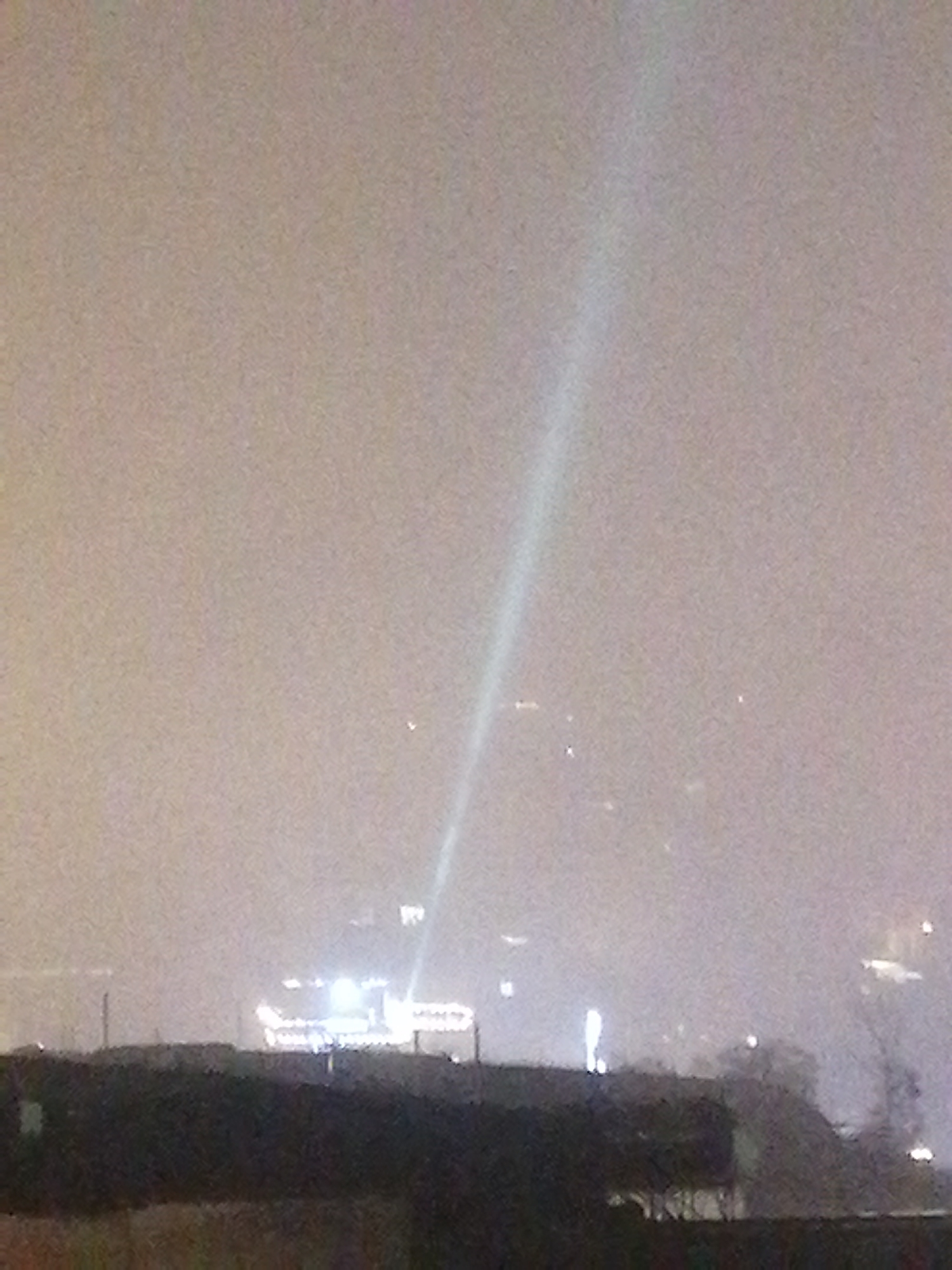 The image shows a scene in Sapa, Vietnam, at night, possibly in Sa Pa Ward, during foggy or misty weather. A bright, narrow beam of light shoots upwards into the sky. The lower portion of the view is a dark, low-lying foreground with irregular dark shapes that might be rooftops or foliage. Above this, a building or structure is illuminated with multiple lights, including strings or arrays of lights. Other, less distinct lights are scattered in the distance.