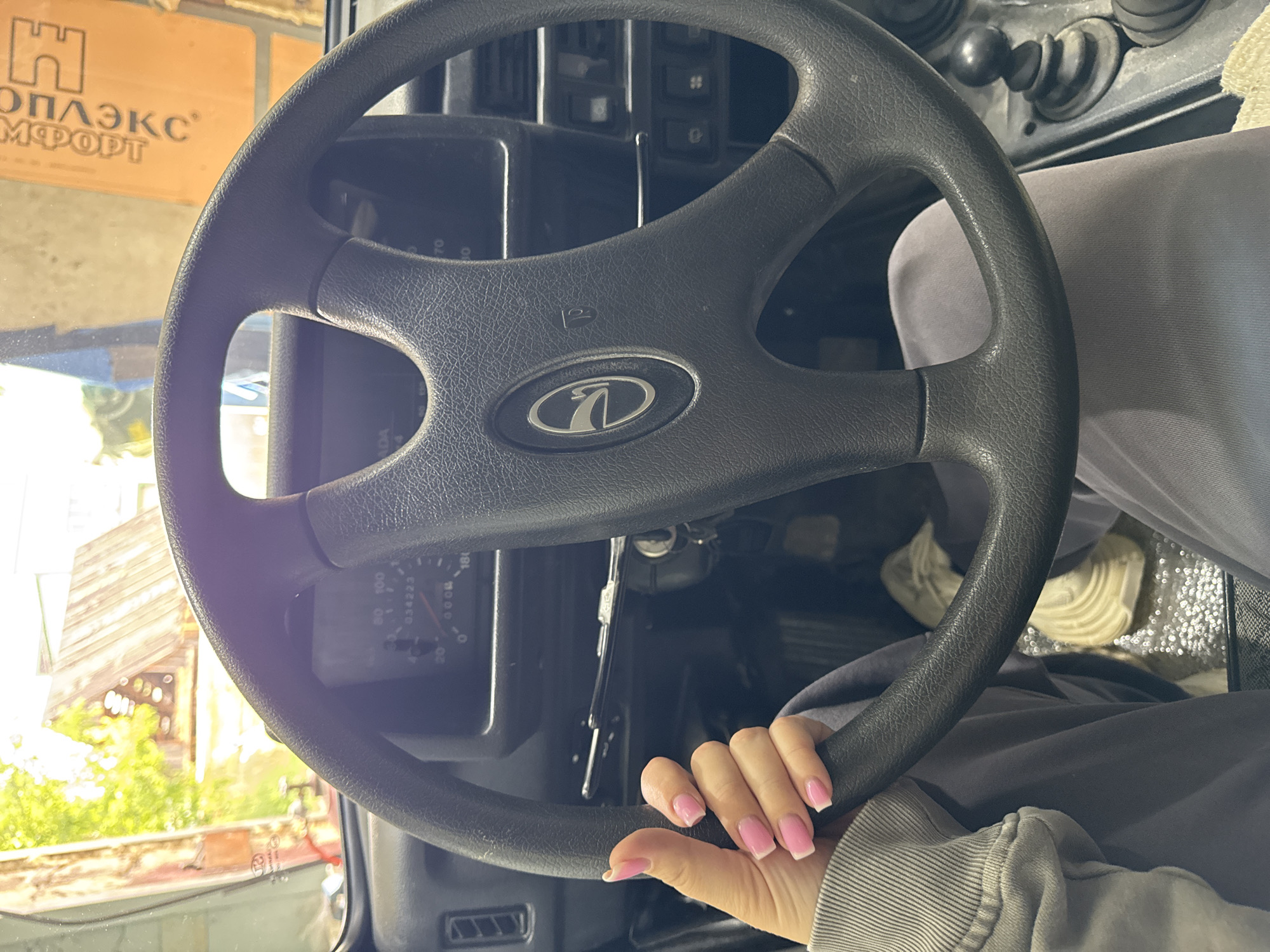The image shows a close-up view of a person's hands on the steering wheel of a car. The person's left hand is resting on the steering wheel, with their thumb extended upwards, showing manicured nails with pink polish and white tips. The car's dashboard is visible behind the steering wheel, including the speedometer and other gauges. The interior of the car appears to be a bit worn. Outside the car, through the windshield, a rustic structure with a wooden roof is visible, along with greenery and a brightly lit, possibly sunny, outdoor environment. The overall setting suggests a rural or semi-rural location. The orange sign in the background, with Russian text, indicates the location is likely in Russia. Specifically, it could be in the city of Горный, Russia, given the available context and common Russian urban signage. The car appears to be parked, possibly in a garage or workshop, given the industrial-looking backdrop in the upper right corner.