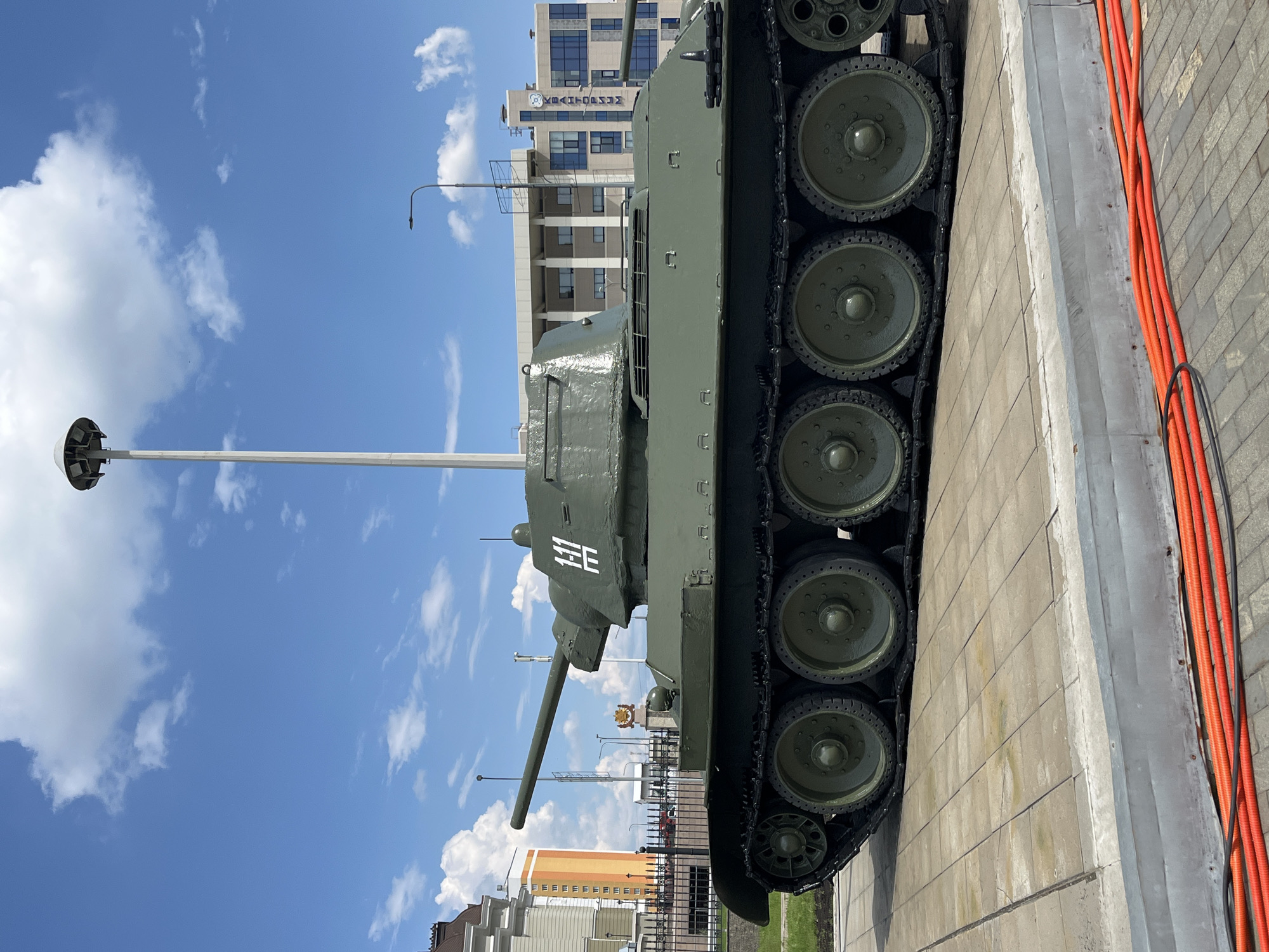 This image captures a T-34 tank, a Soviet-era medium tank, on display outdoors in Verkhnyaya Pyshma, Russia. The tank is painted olive drab and is positioned on a paved area, with its tracks visible. A white emblem with what appears to be Cyrillic characters is painted on the turret. In the background, several modern buildings are visible, including one with the word "KRATOVM" on its facade, suggesting an urban setting. The sky is blue with scattered white clouds, and a tall lamppost stands prominently near the tank. The foreground shows red corrugated tubes lying on the pavement. The scene appears to be a sunny day, possibly in a park or museum dedicated to military history.