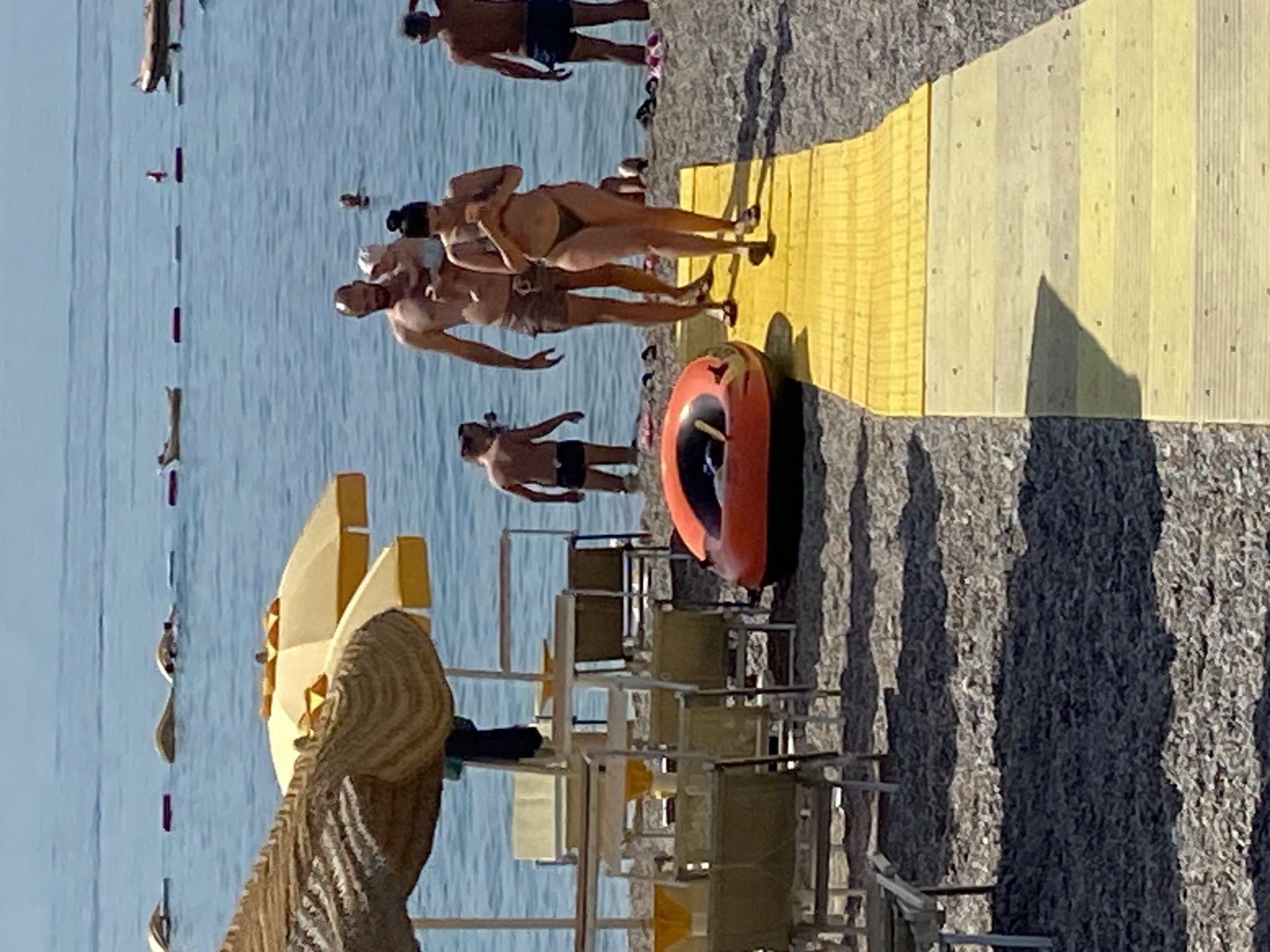 The image depicts a beach scene in Prijevor, Montenegro, on a sunny day. In the foreground, a yellow wooden walkway is laid on a pebble beach. To the left of the walkway, several empty beach chairs and umbrellas are visible. Further back, several people are present. A man carrying a baby walks along the walkway towards the sea. A woman in a bikini walks ahead of him on the walkway, looking at her phone. In the water, a few people are swimming and there are several buoys in the sea. An orange inflatable raft is on the beach near the walkway. Other beachgoers are visible further down the beach. The sea is calm and blue under a clear sky.