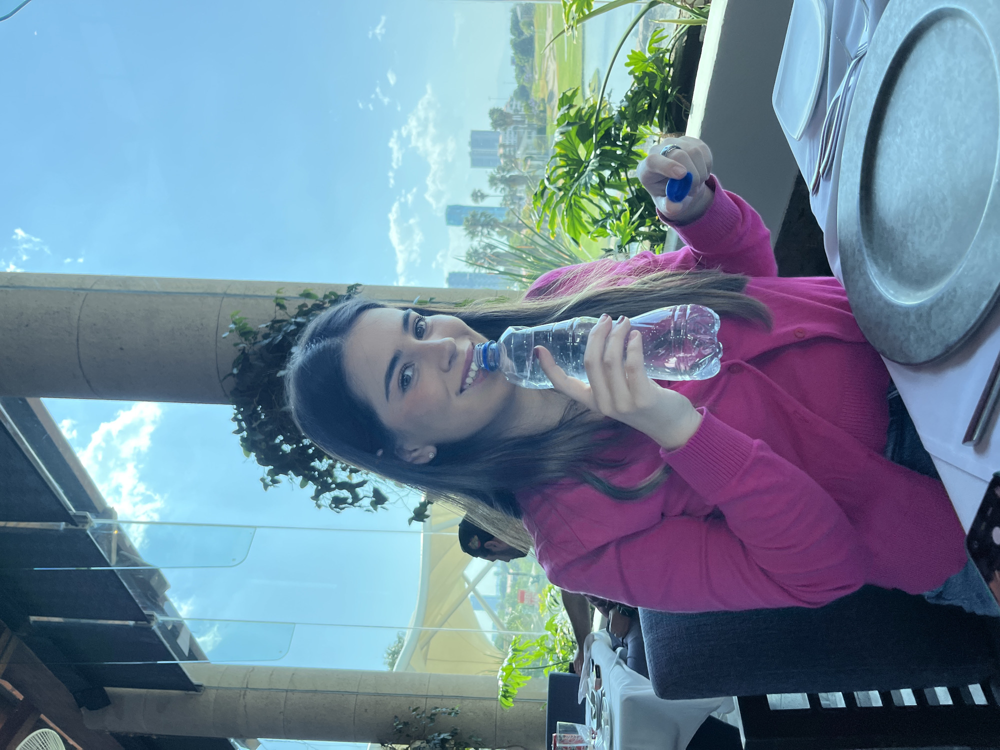 A young adult female with long dark brown hair, wearing a bright pink long-sleeved cardigan, is seated at a white-clothed table inside a restaurant in San Bernardino Tlaxcalancingo, Mexico. She holds a transparent plastic water bottle with a blue cap to her mouth with her left hand, while her right hand holds the detached blue bottle cap. She is smiling and looking directly at the camera. A silver ring is visible on her left ring finger.
The table in the foreground is set with a dark grey circular charger plate, a white rectangular plate, and silverware (fork and knife) to the right. A dark-colored mobile phone rests on the table in the lower left. The woman is seated in a dark-colored upholstered chair with a slatted back. In the background, to the left, the back of a dark-haired male's head and shoulder are visible as he sits at another white-clothed table.
The restaurant features large, floor-to-ceiling glass panels offering an expansive view of the exterior. Visible through these windows is a bright blue sky with scattered white clouds, lush green vegetation including leafy plants and palm trees, a body of water resembling a lake or pond, and manicured green areas indicative of a golf course. In the distant background, multiple modern high-rise buildings form a city skyline, consistent with an urbanized area near San Bernardino Tlaxcalancingo, Mexico. Light-colored architectural columns and dark overhead structures are part of the building's interior. Vines are visible trailing down from overhead structures. A light-colored fabric canopy structure is also visible outdoors, through the window.