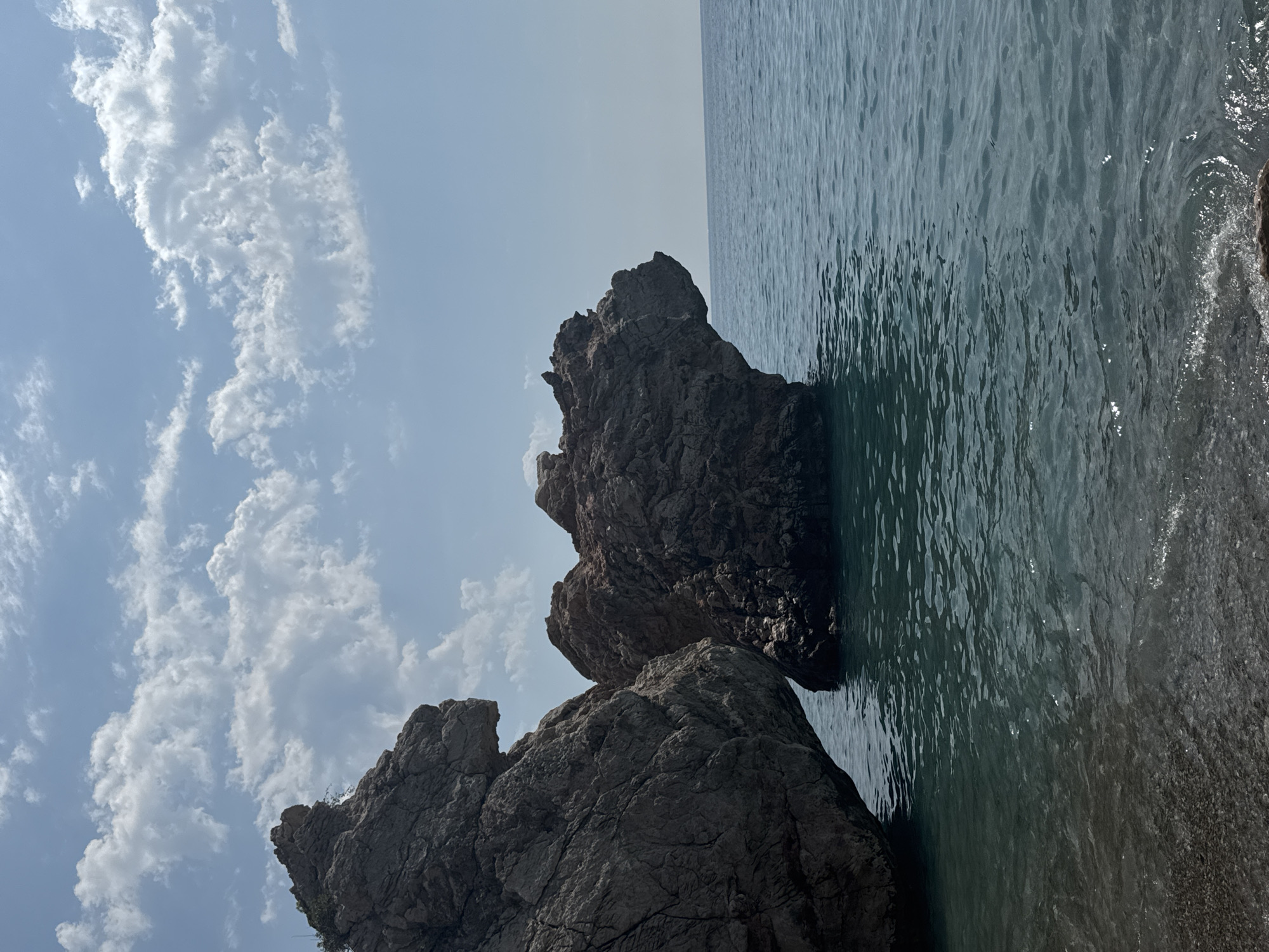 The image shows a rocky coastline with large rock formations jutting out into the clear blue sea. The rocks are dark gray and appear to be heavily weathered, with rough textures and sharp edges. The water is a deep turquoise color, with gentle waves lapping at the shore. The sky above is a pale blue with scattered white clouds. The scene depicts a natural landscape with no visible human presence. This location is likely the coastline of Kemer, Turkey, known for its dramatic rock formations and clear waters.