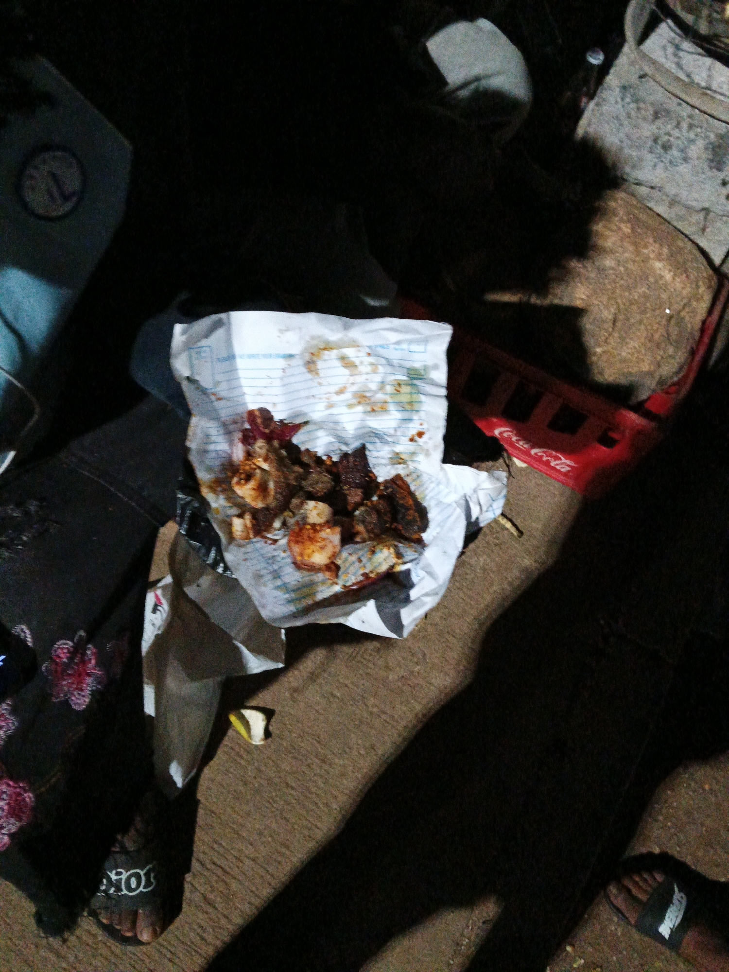 The image depicts an outdoor scene at night, likely in Bukuru, Nigeria, with a focus on discarded food. In the center, a crumpled white paper wrapper, possibly from a fast-food establishment, is filled with pieces of fried meat or chicken, some appearing to be garnished with spices. The food is spread out and partially eaten. To the right of the food, a red Coca-Cola crate is visible, suggesting a commercial or public setting. The ground appears to be paved or a dirt pathway. Shadows obscure much of the background, indicating low lighting conditions. On the left, a person's leg wearing a black slipper with white lettering is visible, partially in shadow. Another foot wearing a black slipper with white lettering is visible in the lower right corner of the image. A large, dark shadow dominates the right side of the image, possibly cast by a person or object out of frame. The overall impression is that of a casual, possibly late-night gathering or an area where food has been consumed and discarded.