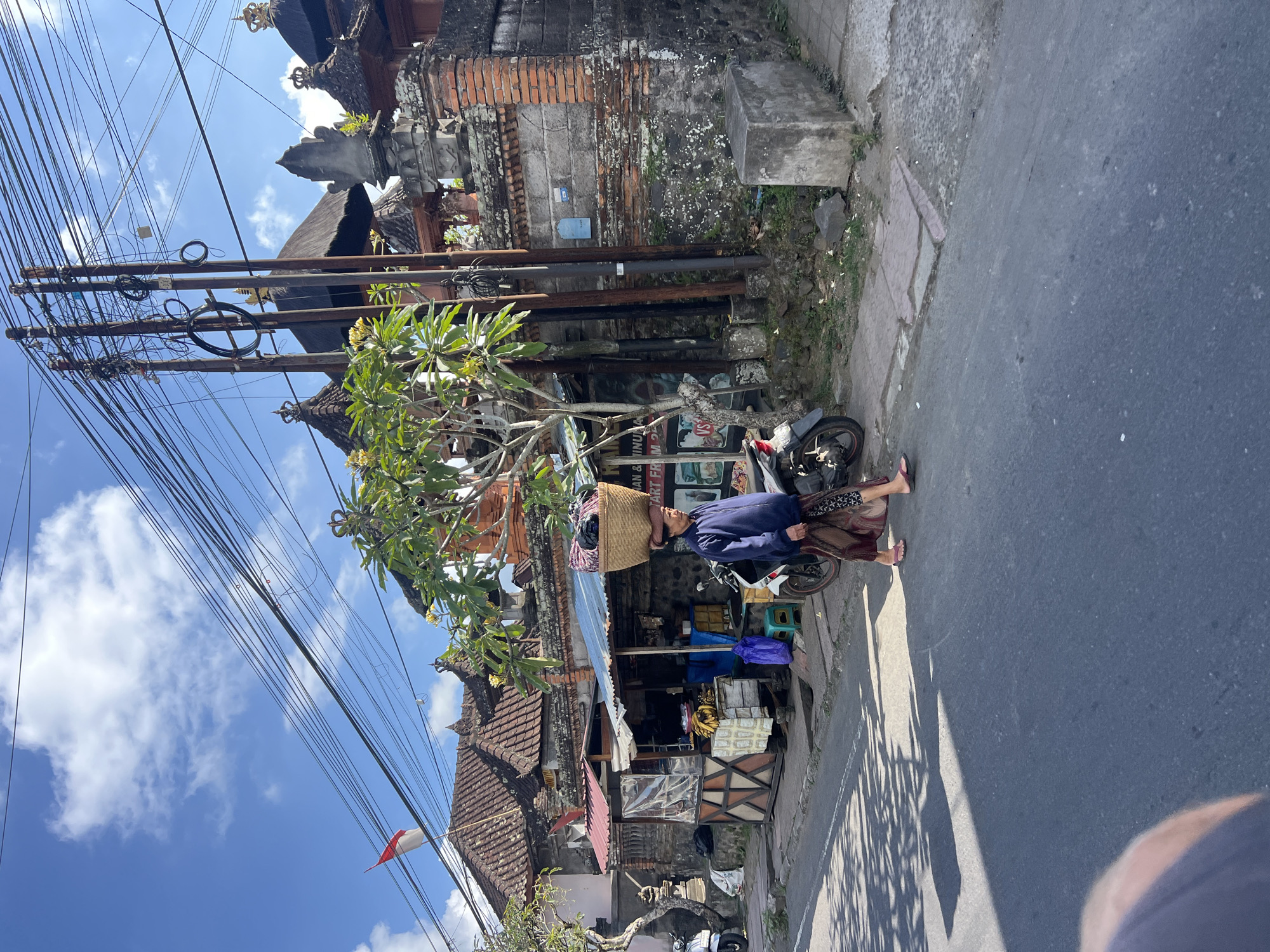In Tampaksiring, Indonesia, a woman walks along a paved street, carrying a woven basket filled with items on her head. She wears a blue long-sleeved shirt and a patterned sarong. She is walking past a small shop with a blue tarp roof, where bananas and other goods are displayed. A motorcycle is parked next to the shop. Behind the shop, a traditional Balinese building with a tiled roof and ornate decorations is visible. Power lines are strung across the sky, supported by wooden poles. The sky is blue with scattered white clouds. The sun casts shadows on the street.