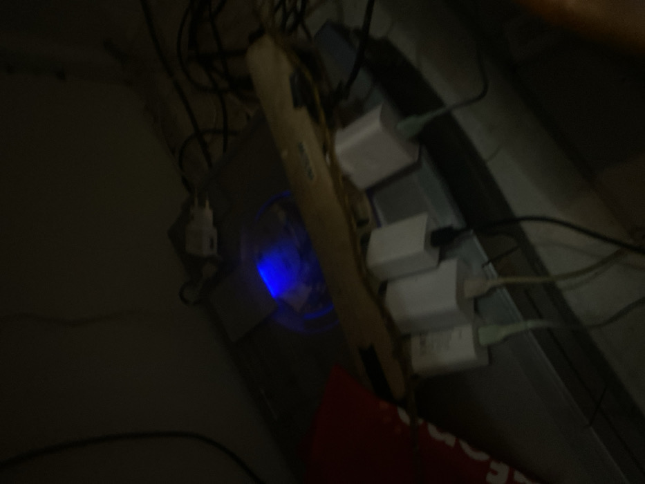The image depicts a dimly lit indoor scene, specifically an area with electronic equipment, located in Cimenyan, Indonesia. No individuals are present in the frame.
In the center, a dark, rectangular electronic device, possibly a computer tower or game console, exhibits a prominent internal blue light, likely from a cooling fan or indicator. To the immediate right of this device, a beige or light-colored power strip is positioned horizontally, resting on a surface. Four white, rectangular power adapters/chargers are plugged into this power strip. Multiple cables extend from these adapters; at least two light green cables and one off-white cable are visible originating from the bottom of the chargers, leading downwards and to the right, connecting to unseen devices. Numerous dark cables are tangled behind and around the primary electronic device and power strip, indicating a cluttered setup.
On the far left, a portion of a red object, possibly fabric or a bag, is visible. The background consists of a plain, light-colored wall or surface. The overall environment appears to be a corner or desk area dedicated to electronic components and their associated power connections.