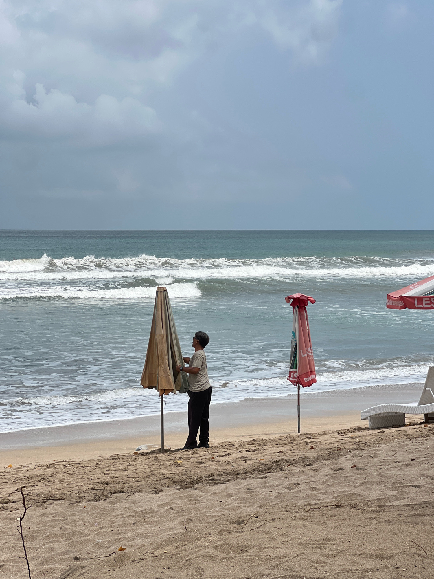 A person is standing on a sandy beach in Kuta, Indonesia, closing a large, tan-colored beach umbrella. The umbrella is partially furled and leaning. To the right of this umbrella is a closed, red and white striped umbrella, with text visible on it that includes "LES". Further to the right, a white lounge chair is partially visible. The ocean is in the background with moderate waves crashing on the shore. The sky is overcast with gray clouds. The overall atmosphere is calm and serene.