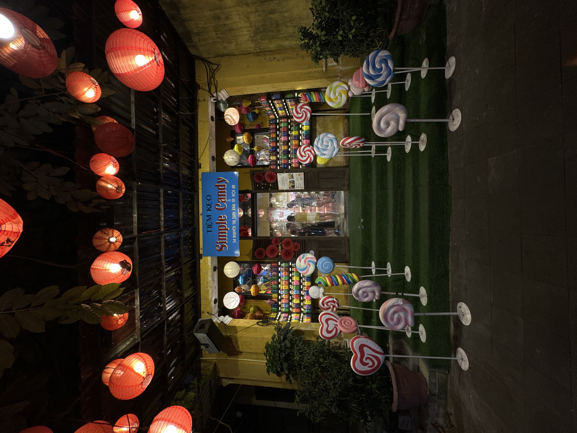 A brightly lit candy store in Hoi An, Vietnam, is showcased at night. The store, with its yellow facade and covered entrance, is adorned with numerous red lanterns hanging from the ceiling, creating a festive and warm atmosphere. In front of the store, a collection of oversized lollipop and heart-shaped candy decorations stands on white poles, arranged on a green carpeted walkway leading up to the entrance. Inside, shelves are stocked with jars of colorful candies, visible through the open doorway. Several people are seen browsing inside the store. The scene is a testament to the vibrant and charming commercial scene of Hoi An.