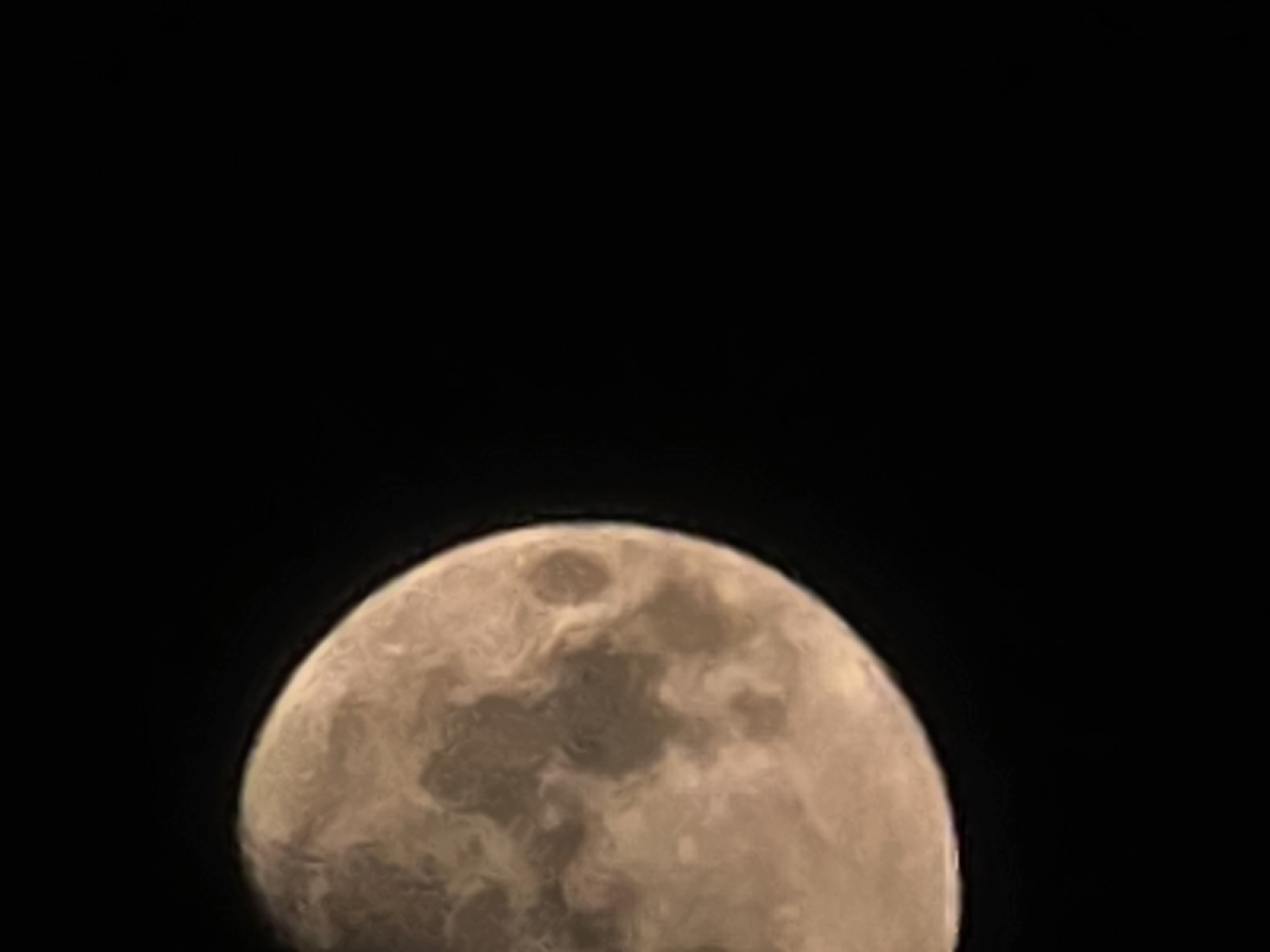 The image is a close-up, slightly blurry photograph of the moon against a black background. The moon is partially illuminated, with a significant portion of its surface visible. The surface shows discernible variations in light and shadow, suggesting the presence of craters and other geological features, though these are not sharply defined due to the image quality. The color of the moon appears to be a warm, light brown or tan. The composition is focused on the moon, with no other objects or elements present in the frame. The photograph does not depict any urban environment, and therefore no specific location in the city of Yambol, Bulgaria, can be identified.