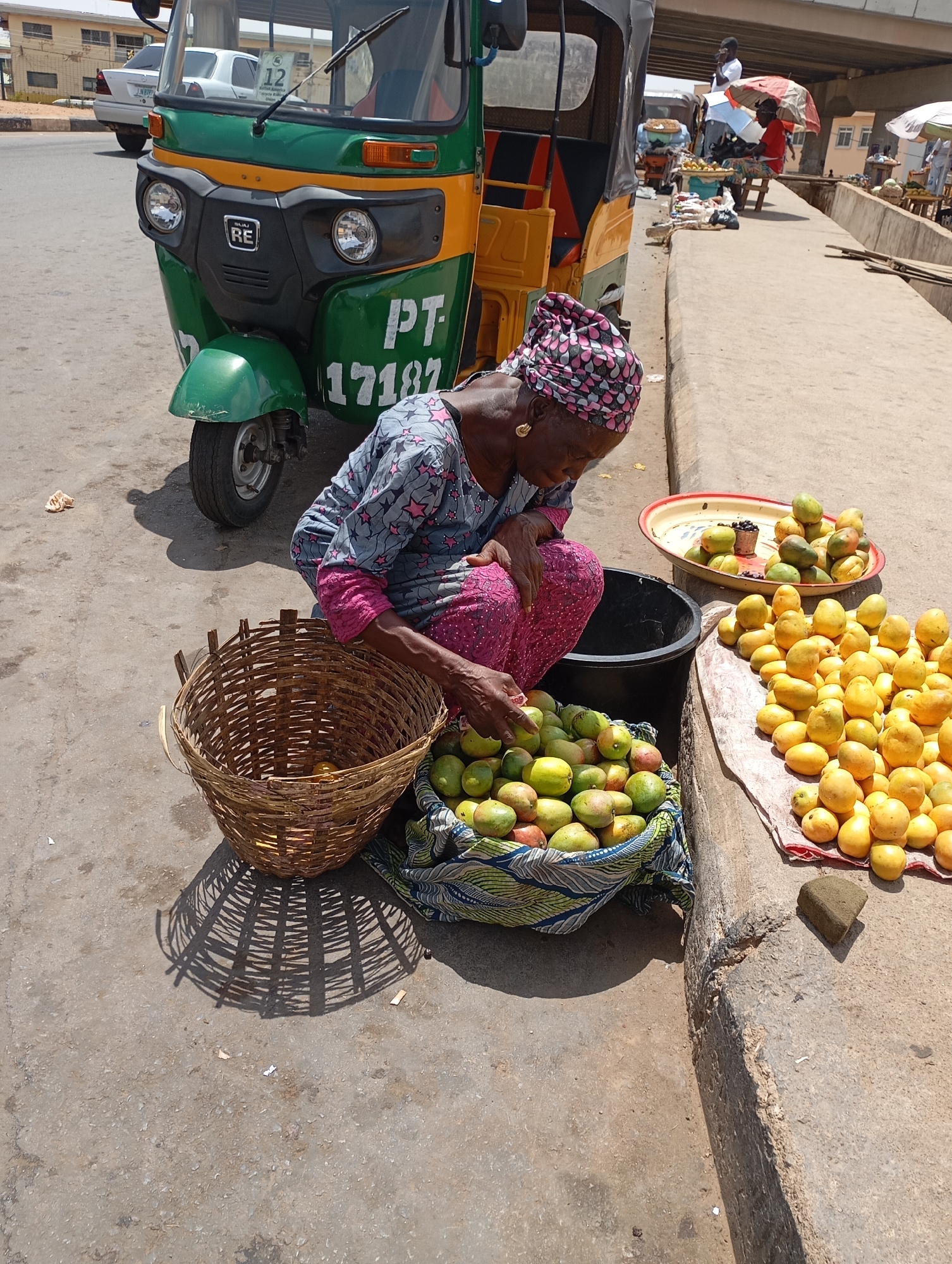 The image shows a street scene in Jos, Nigeria. An elderly woman wearing a patterned headwrap and a star-patterned top and pink pants is crouching next to a curb. She is sorting mangoes from a fabric-lined basket into a black plastic container. To her left is a woven basket, and to her right, mangoes are arranged on a cloth and in a metal tray. Behind her is a green and yellow auto-rickshaw with the registration number "PT 17182." In the background, people are walking on a raised platform or bridge, some under umbrellas.