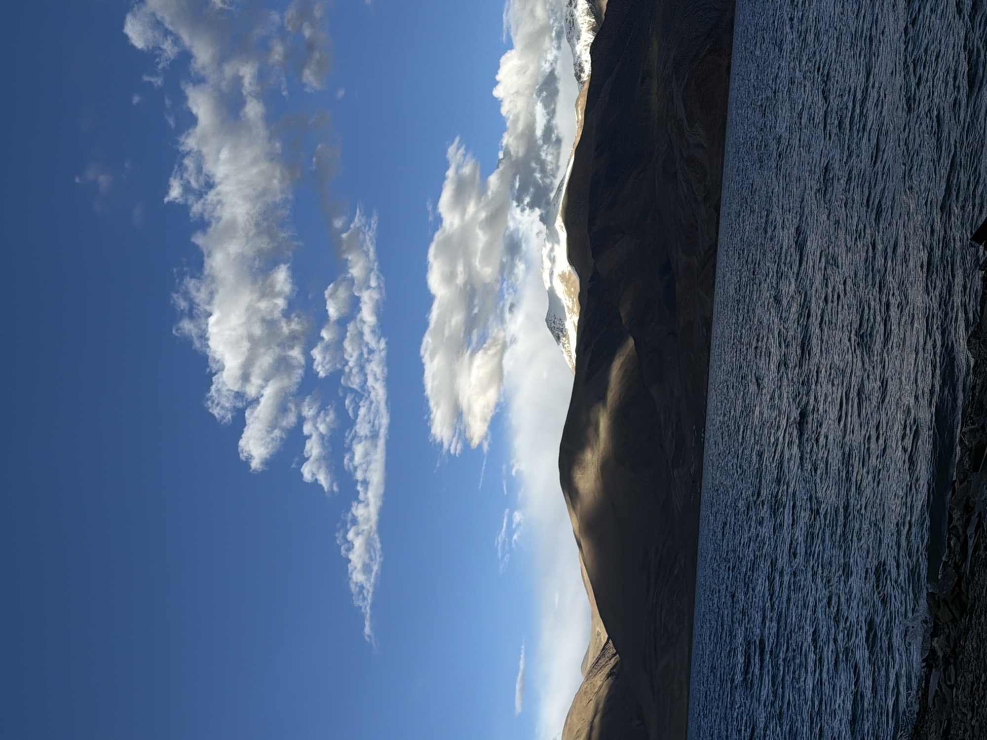 The image depicts a vast, serene lake in a mountainous region, likely in India, under a bright blue sky dotted with white, fluffy clouds. The mountains in the background are rugged and brown, with some peaks capped in snow. Sunlight casts dramatic shadows across the slopes, creating a play of light and dark. The water of the lake is a deep blue, with ripples on the surface suggesting a gentle breeze. The shoreline is rocky and sparsely vegetated. The overall scene is one of natural beauty and tranquility, conveying a sense of remoteness and awe.