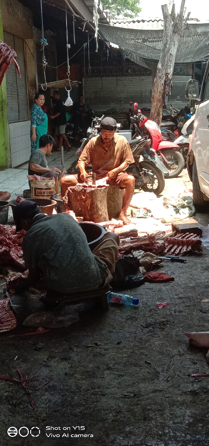 The scene depicts an active, open-air butchery operation located in the city of Maron, Indonesia. The environment features an uneven, likely concrete or dirt ground, partially shaded by a makeshift tarpaulin roof and corrugated metal structures. A dead tree trunk stands vertically in the mid-ground right. Overhead, ropes with metal hooks are suspended from a beam.
In the foreground, a male individual wearing a grey t-shirt, light-colored pants, and a dark cap is seated, facing away from the camera, hunched over a large, dark metal basin, appearing to process meat. Adjacent to him, a plastic bottle of Mizone is on the ground.
In the mid-ground, a second male individual, barefoot, wearing a brown t-shirt and brown shorts with a black cap, is seated on a low stool. He is actively butchering raw meat with a cleaver on a wooden chopping block. Various cuts of raw meat, including rib cages and bones, are scattered on the ground around him, alongside what appear to be bloodstains.
Further in the mid-ground left, another male individual in a green t-shirt is seated, smoking a cigarette, observing the activity. To his left, a female individual in a patterned blue/green dress and a light green apron stands, looking towards the left side of the frame.
In the background, several motorcycles of various models (e.g., red and black scooters) are parked, and parts of a white van are visible on the far right. Several indistinct figures are present in the background, near the parked vehicles and structures. The lighting suggests a bright, sunny day.