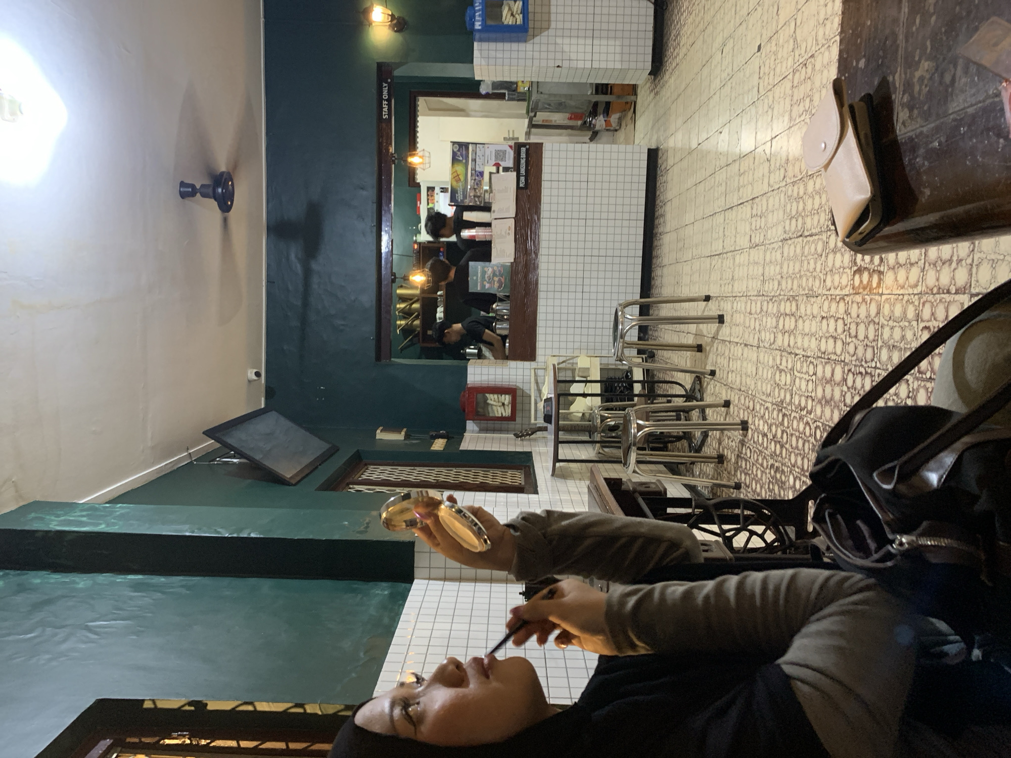 A woman wearing a black headscarf and gray sweater applies makeup with a brush while seated inside a brightly lit restaurant, presumably in Tamanbaru, Indonesia. The restaurant has a green wall and a ceiling fan. A counter with staff is visible in the background, along with tables and chairs. A small makeup compact is in the woman's hand. Tiled floors and walls are also present.