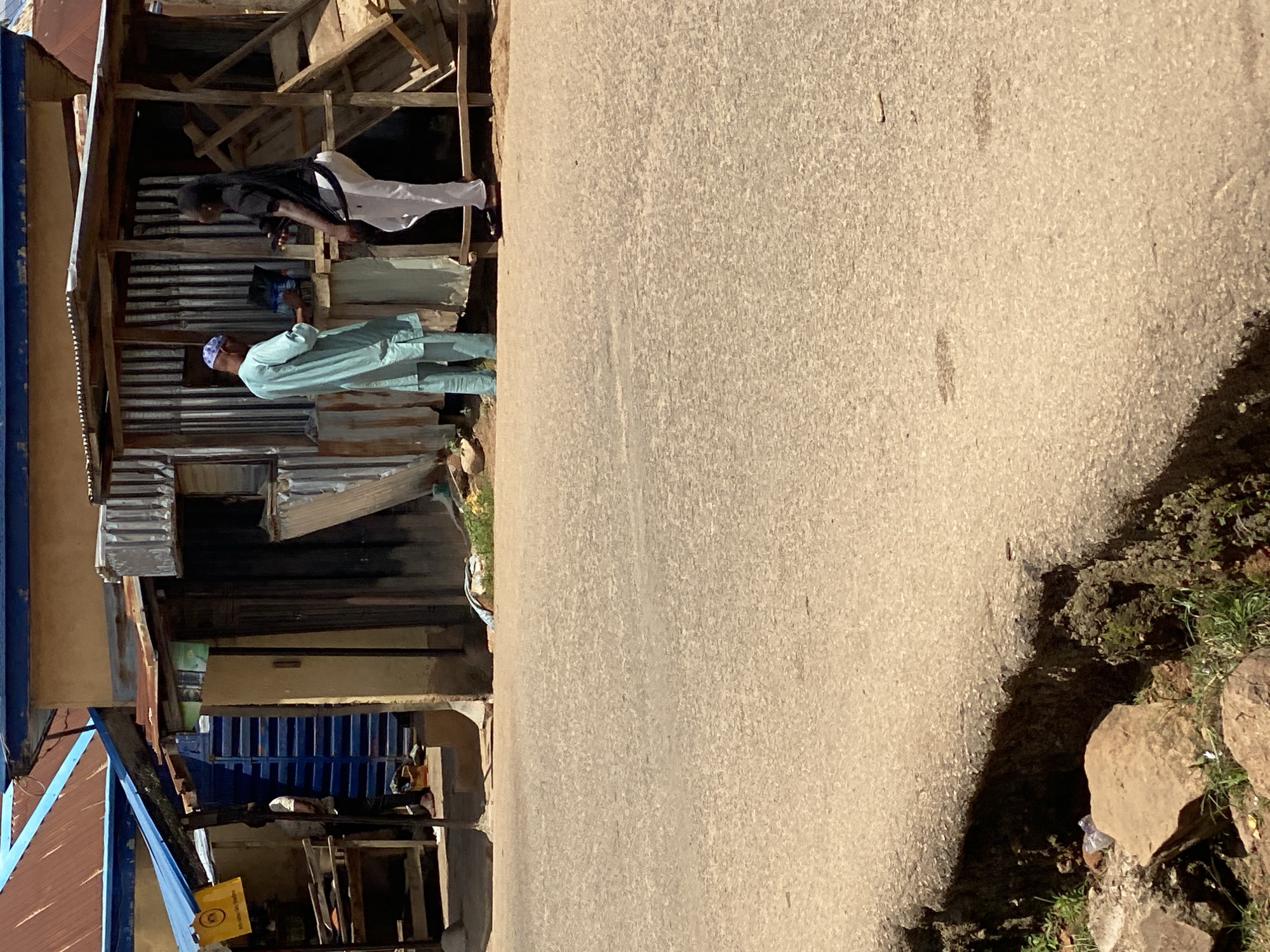 The image depicts an outdoor scene in Jos, Nigeria, featuring a modest, open-air stall constructed from corrugated metal and wooden planks. Two individuals are present at the stall. On the left, a man wearing a light blue kaftan and a matching cap stands facing a counter, seemingly engaged in a transaction or conversation. To his right, a woman wearing a black t-shirt and white trousers stands behind the counter, holding a black item, possibly a phone or camera. The stall is situated on a dirt road, with a visible drainage ditch on the left side of the foreground. Various items are visible within and around the stall, including what appears to be stacked goods and wooden shelving. The overall setting suggests a typical marketplace or roadside vendor location in Jos.