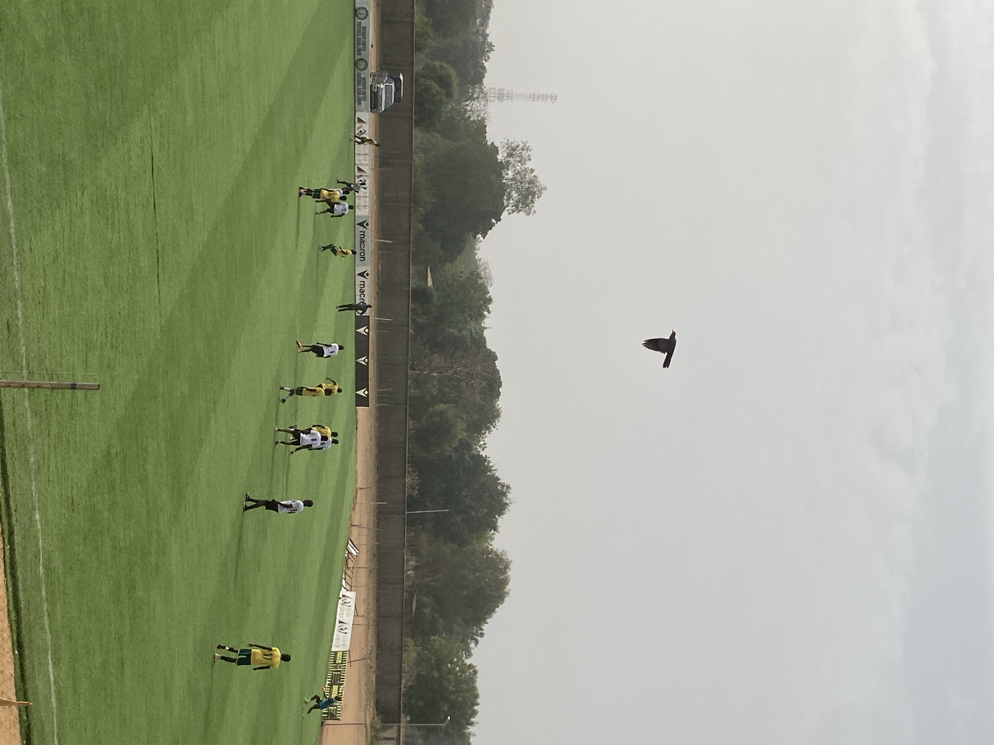 The image shows a soccer field in Tudun Wada, Nigeria. Players in yellow and white uniforms practice on the green field, adjacent to a wall and some trees. A single bird flies in the hazy sky. A tall communication tower is visible in the distance behind the trees.