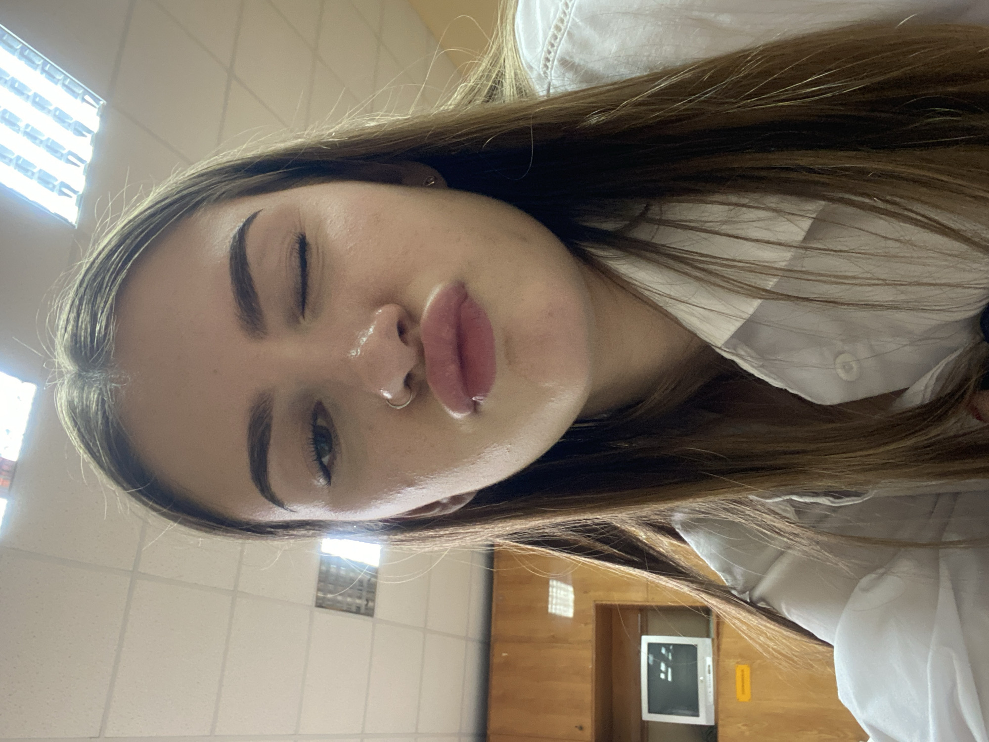 A young woman is pictured in a close-up, selfie-style shot, indoors. She has long, brown hair that is parted in the middle and falls around her shoulders. Her right eye is open, while her left eye is closed in a wink. Her lips are pursed in a kiss-like gesture, and she has a nose ring. She is wearing a white collared shirt. In the background, there are wooden cabinets and a white computer monitor on a shelf. Overhead fluorescent lighting is visible, typical of an indoor space such as a classroom or office. The overall impression is of a casual, personal photograph.