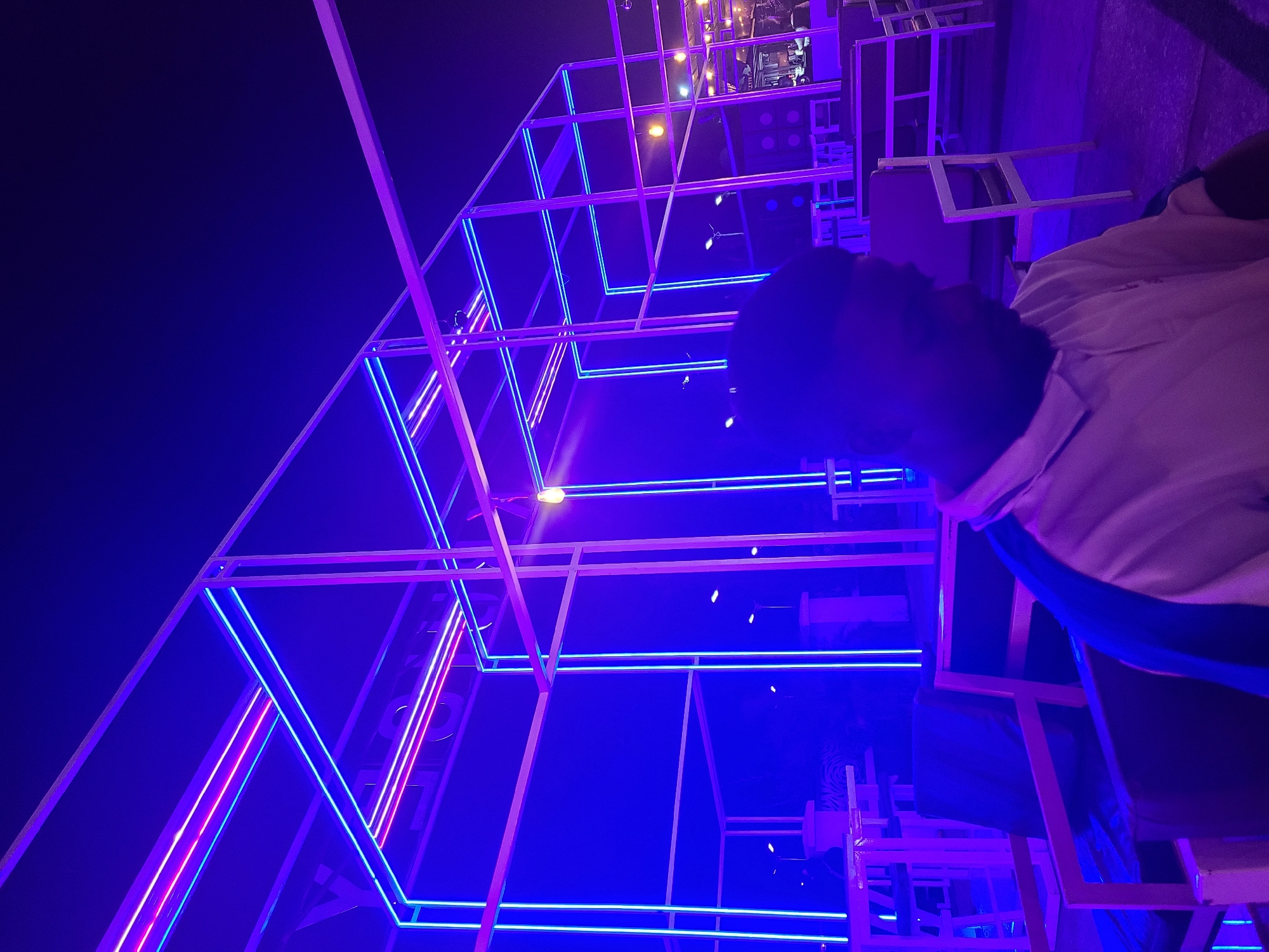 The image depicts a person in the right foreground, partially visible, wearing a white shirt with blue trim. They are seated in an area with geometric, box-like structures outlined by illuminated linear lights in shades of blue and pink. These structures appear to be part of an architectural or decorative installation. In the background, more of these structures and ambient lighting are visible. The lighting suggests it is nighttime or a dimly lit interior. Based on the prompt instructions, this scene is located in the city of Gura Top, Nigeria.