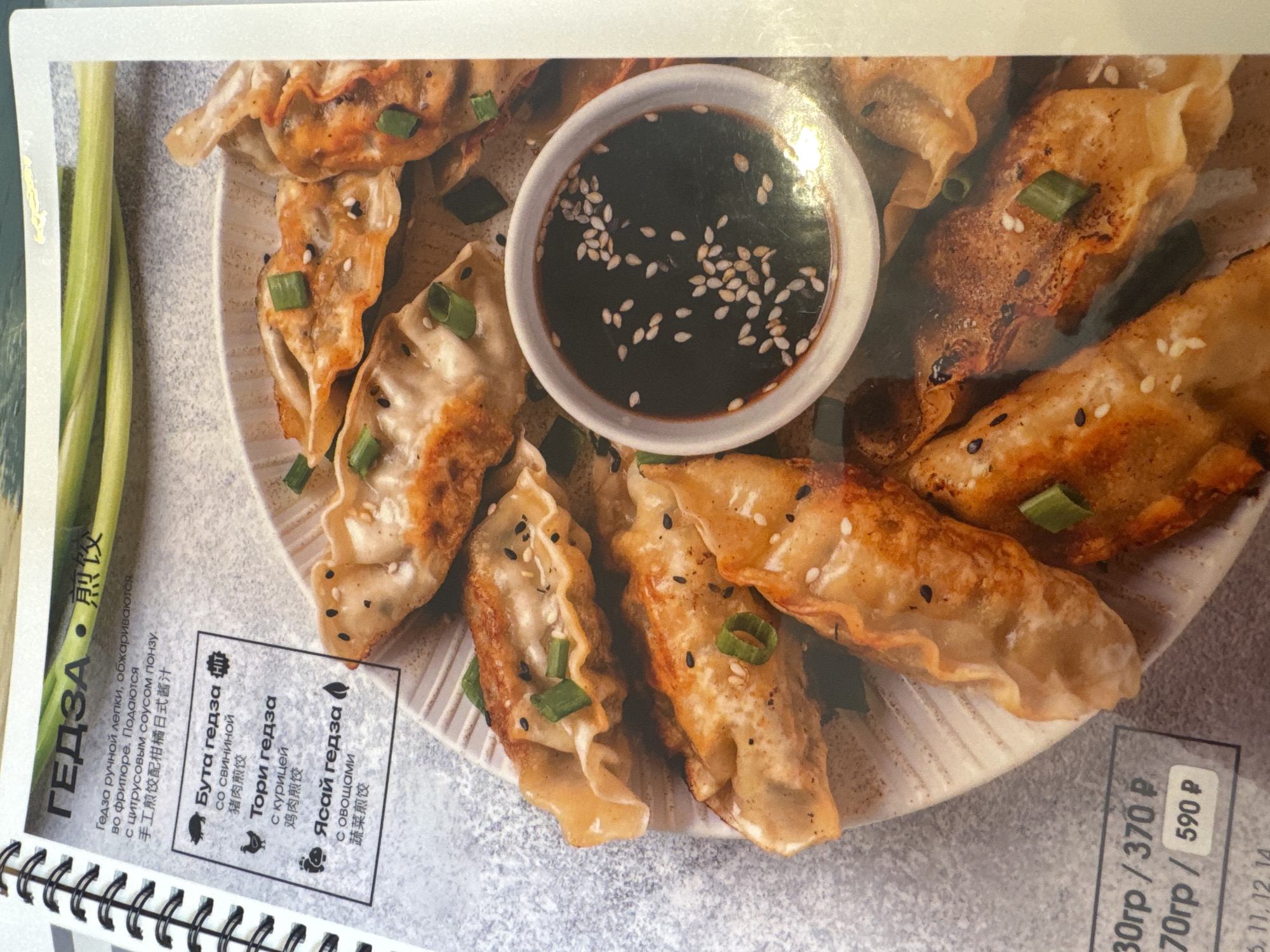 This image displays a close-up of a menu page featuring a dish identified as "ГЕДЗА" (Gedza), which are fried dumplings. The central focus is a white plate filled with numerous golden-brown dumplings, lightly pan-fried or deep-fried, and garnished with chopped green onions and black and white sesame seeds. A small white bowl containing a dark dipping sauce, also topped with sesame seeds, is prominently placed on the plate.
The surrounding menu text, presented in both Russian and Chinese characters, provides details about the dish. The Russian description states, "Гедза ручной лепки, обжариваются во фритюре. Подаются с цитрусовым соусом понзу." (Hand-sculpted gedza, deep-fried. Served with citrus ponzu sauce.) The Chinese characters "煎饺" confirm "fried dumplings," and the more detailed Chinese text translates to "Hand-fried dumplings with citrus Japanese sauce."
Three varieties of Gedza are listed with corresponding icons:
1. "Бута гедза" (Buta gedza, with a pig icon) "со свининой" (with pork).
2. "Тори гедза" (Tori gedza, with a chicken icon) "с курицей" (with chicken).
3. "Ясай гедза" (Yasai gedza, with a leaf icon) "с овощами" (with vegetables).
Pricing is visible at the bottom right: "30гр/ 370 р" (30 grams / 370 rubles) and "70гр / 590 р" (70 grams / 590 rubles). The image suggests a restaurant setting in Saint Petersburg, Russia, given the language and context. No people are visible; the media is a food advertisement or menu item photograph.