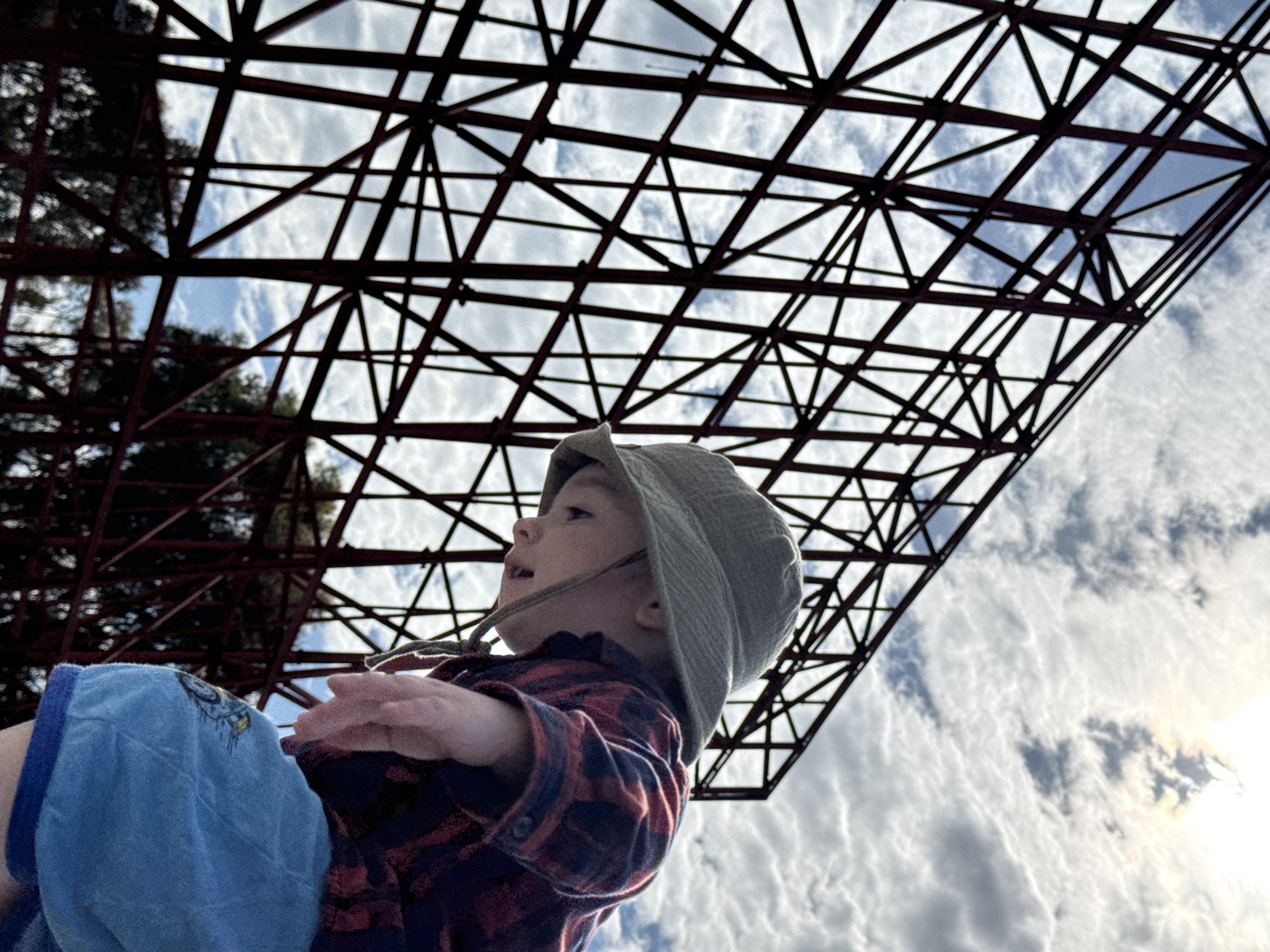 A low-angle shot features a toddler wearing a green bucket hat and a red and black plaid shirt, gazing upwards. The child is partially obscured by a blue object, possibly clothing. Above the child is a dense structure of intersecting metal beams against a backdrop of a cloudy sky, with the sun partially visible on the right. The location appears to be in an outdoor park or recreational area in the city of Sirius, Russia.