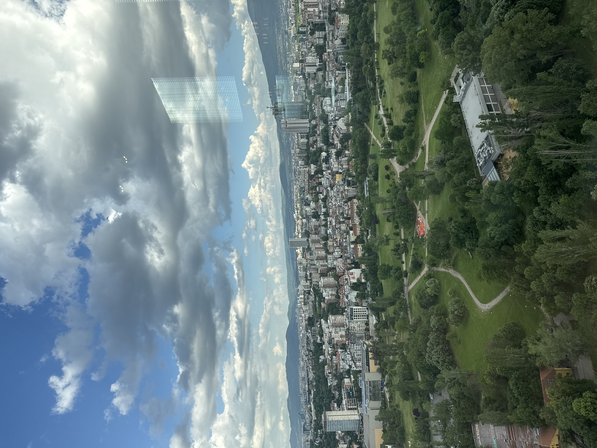 The image shows an aerial view of Sofia, Bulgaria, partially obscured by reflections in a window.
On the right, a green park stretches out with pathways winding through the trees. Buildings are interspersed among the trees in the park.
Adjacent to the park, a dense urban area extends towards the center of the image. The cityscape is dominated by a mix of residential and commercial buildings of varying heights. A mountain range is visible in the distance, partially obscured by clouds.
The sky is filled with a mix of blue patches and grey, heavy clouds. In the upper-left, the reflection of another building is visible in the window.
