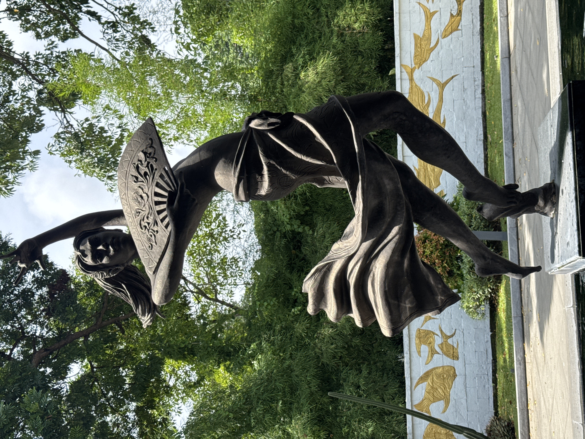 A bronze statue of a woman is located outdoors. She has a fan in her hand, and wears a garment that exposes her midriff. The statue has been identified as a sculpture of Garuda Wisnu Kencana Cultural Park in the city of Kuta Selatan, Indonesia. The statue is situated on a tiled platform decorated with images of golden fish. Trees are behind the sculpture.