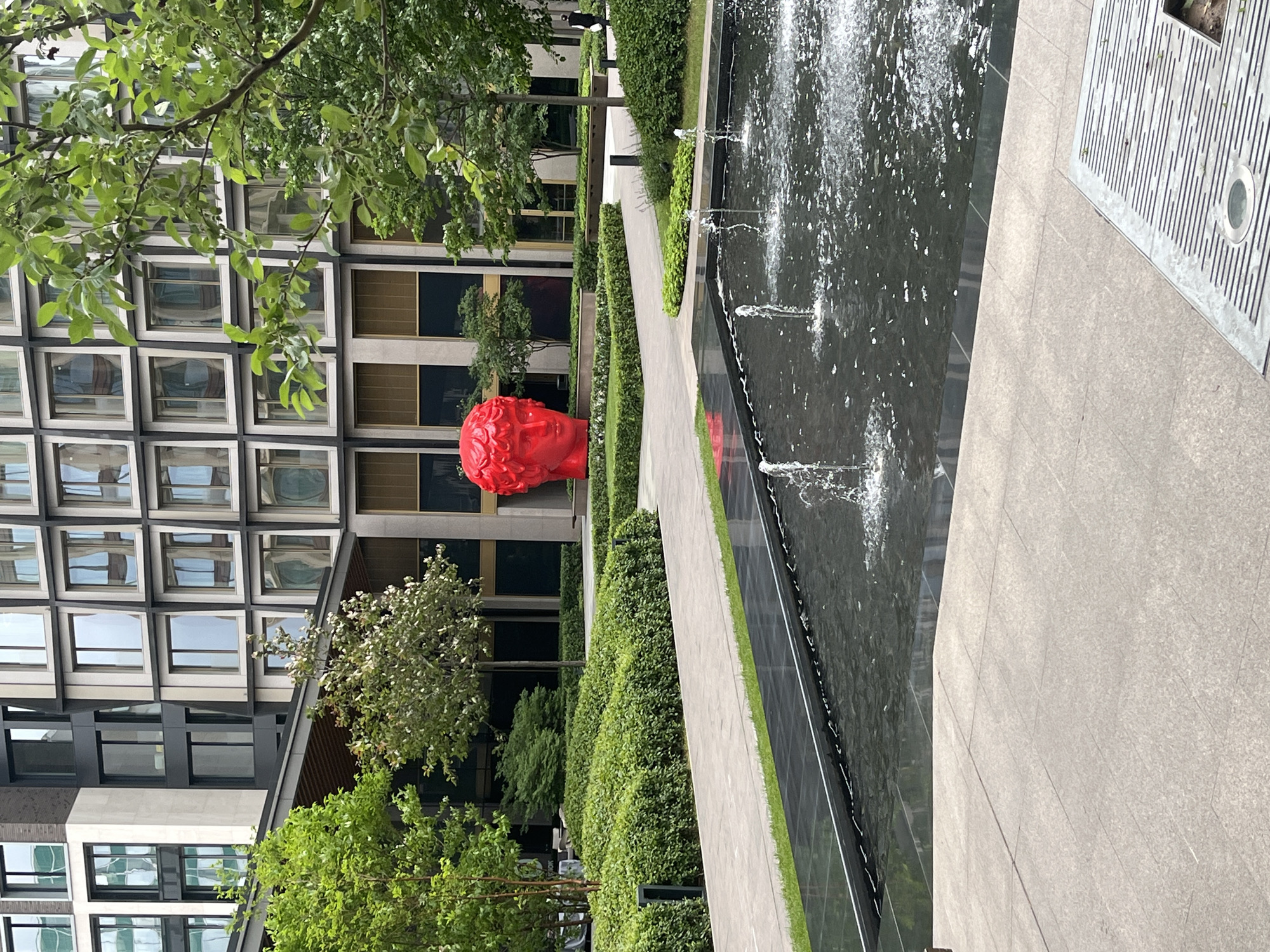 The image shows an outdoor plaza with a modern building in the background and a fountain in the foreground. The building has a grid-like facade with numerous windows. Lush green landscaping, including manicured hedges and trees, surrounds a paved walkway that leads to the building. A large, bright red sculpture of a human head is placed prominently in the landscape. The fountain in the foreground is rectangular with water jets spraying upwards. The plaza is paved with large, light-colored tiles. A single person, dressed in dark clothing, is walking away from the camera on the right side of the frame. The overall impression is one of contemporary urban design with a touch of artistic flair.