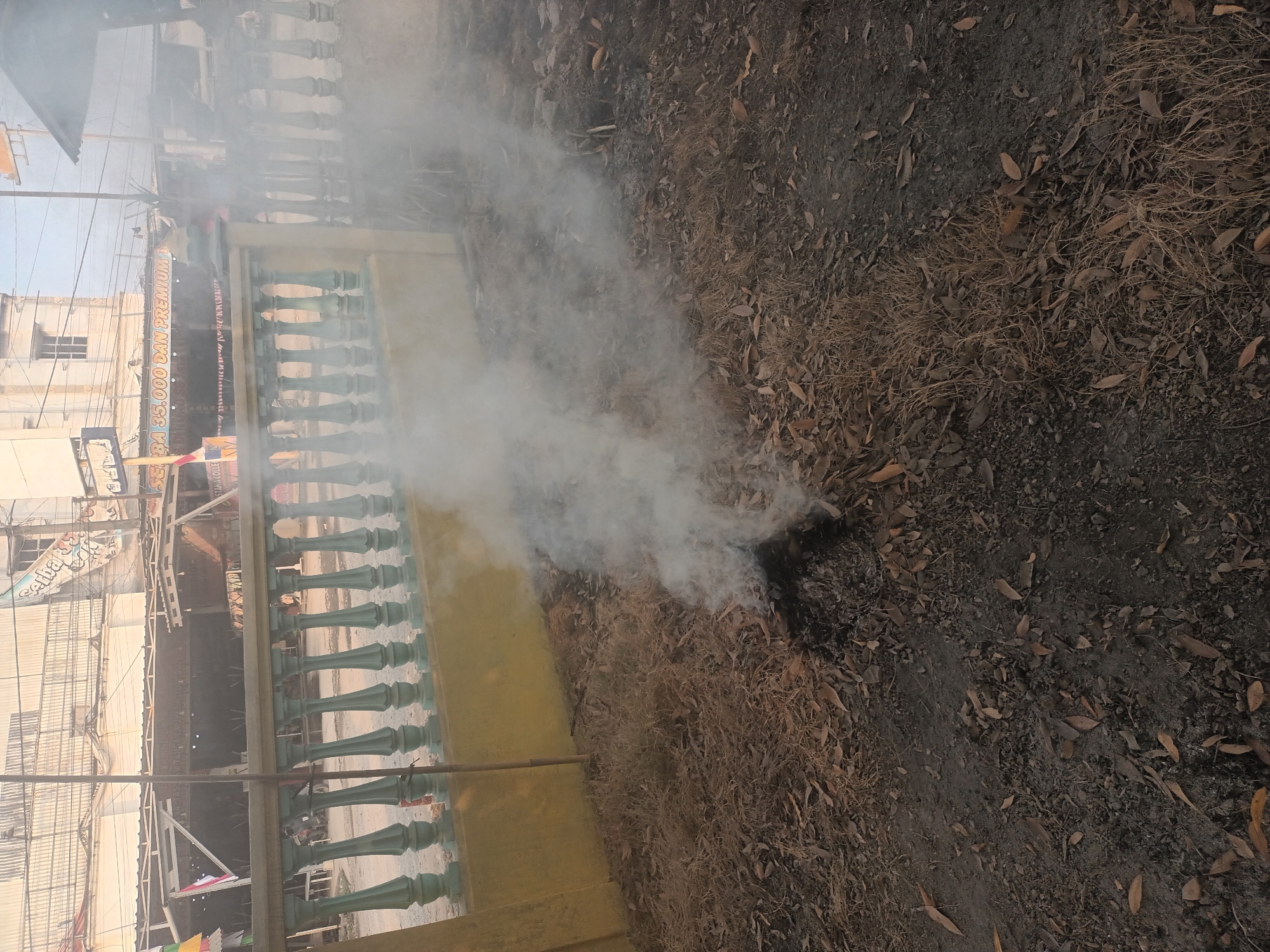 In Mulyo Asri, Indonesia, a patch of dry leaves and twigs smolders on the ground, emitting a plume of white smoke that drifts to the right. The fire is located in the foreground, with charred remnants visible beneath the smoke. Behind the fire, a low, light green balustrade with cream-colored posts stretches horizontally across the frame. Beyond the balustrade, a building with a light-colored facade and multiple windows stands, featuring a sign with text in Indonesian. The sky above is clear.