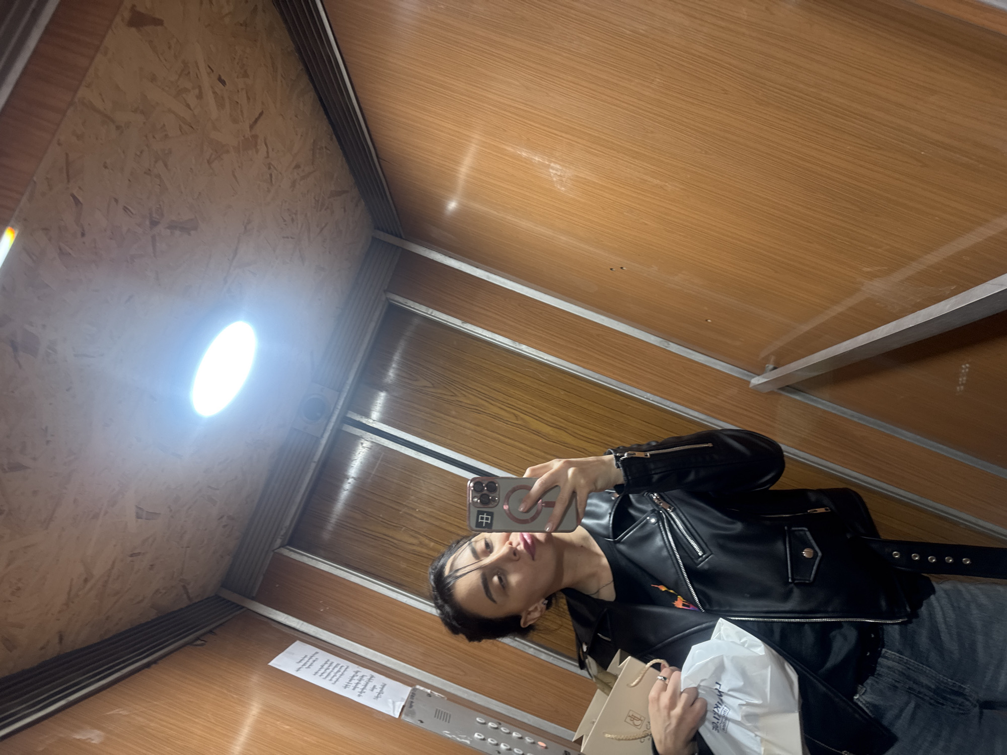A person takes a selfie in an elevator with wooden walls. The elevator appears to be in a building in Tbilisi, Georgia. The person is wearing a black leather jacket and jeans and is holding a phone in their left hand. They are also holding shopping bags in their right hand. There is a light fixture on the ceiling of the elevator, and there is a paper adhered to the wall next to the floor buttons.