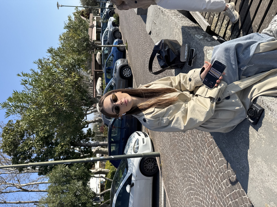 The image depicts an outdoor scene in Sirmione, Italy, under clear daylight conditions. A female individual, approximately 20-30 years of age, is seated on a low, light grey stone or concrete barrier. She has long brown hair with lighter streaks, wears oval-shaped sunglasses with amber lenses, and has a white earbud in her left ear. Her expression is neutral, and she is looking directly at the camera. She is dressed in a light beige trench coat over blue distressed jeans and light-colored sneakers, the latter partially visible on her right foot. In her left hand, she holds a black smartphone with a clear case, its screen displaying an interface. A black compact camera rests on the barrier to her left. To her right, on the same barrier, are a black bucket bag, a patterned glasses case, and a thin object resembling a pen. The lower legs of another person, clad in white pants and light-colored sneakers, are visible to the right, adjacent to wooden planks beneath the barrier.
The immediate foreground features cobblestone pavement to the left of the seated individual. Behind her and extending into the background is a paved parking area with a row of multiple parked cars, including a white Fiat Punto, a dark blue hatchback, and several grey SUVs. Behind the vehicles, a line of trees with green foliage is visible. In the further background, structures resembling buildings with white awnings and light-colored roofs are present under a clear blue sky. A dark grey lamppost stands vertically on the left side of the image. The setting suggests a lakeside or waterfront promenade.