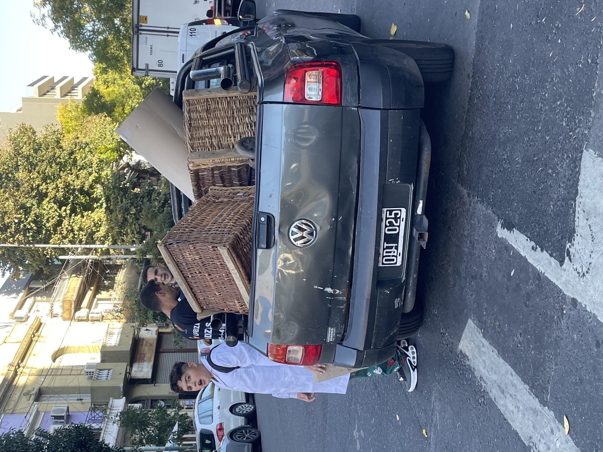 An outdoor street scene in Buenos Aires, Argentina, is captured on a bright, sunny day. The image, originally rotated 90 degrees counter-clockwise, centers on the rear of a dark gray Volkswagen pickup truck, likely a Saveiro model.
The truck's bed is loaded with cargo, most prominently two large, rectangular wicker baskets, one stacked atop the other. Another light-colored, flat object, possibly cardboard or paneling, is also visible in the truck bed. The truck features a Volkswagen emblem on its tailgate and a license plate that reads "OBT 025," consistent with Argentine registration plates.
Two men are positioned near the back of the pickup truck, appearing to handle the wicker baskets, suggesting an activity of loading or unloading. One man, wearing a dark t-shirt with partial text "UBUZA" visible, is on the left, looking towards the other man whose head is partially obscured by the baskets. Standing behind the truck, a young boy or teenager, wearing a white lab coat (a common school uniform in Argentina) over patterned pants and sneakers, looks directly at the camera with a neutral expression.
In the background, a white delivery truck with the numbers "110" and "80" on its side is partially visible. Further down the street, another white car is parked. The setting includes multi-story buildings, some with visible air conditioning units, and lush green trees, indicative of an urban environment. The ground is asphalt with white painted lines.