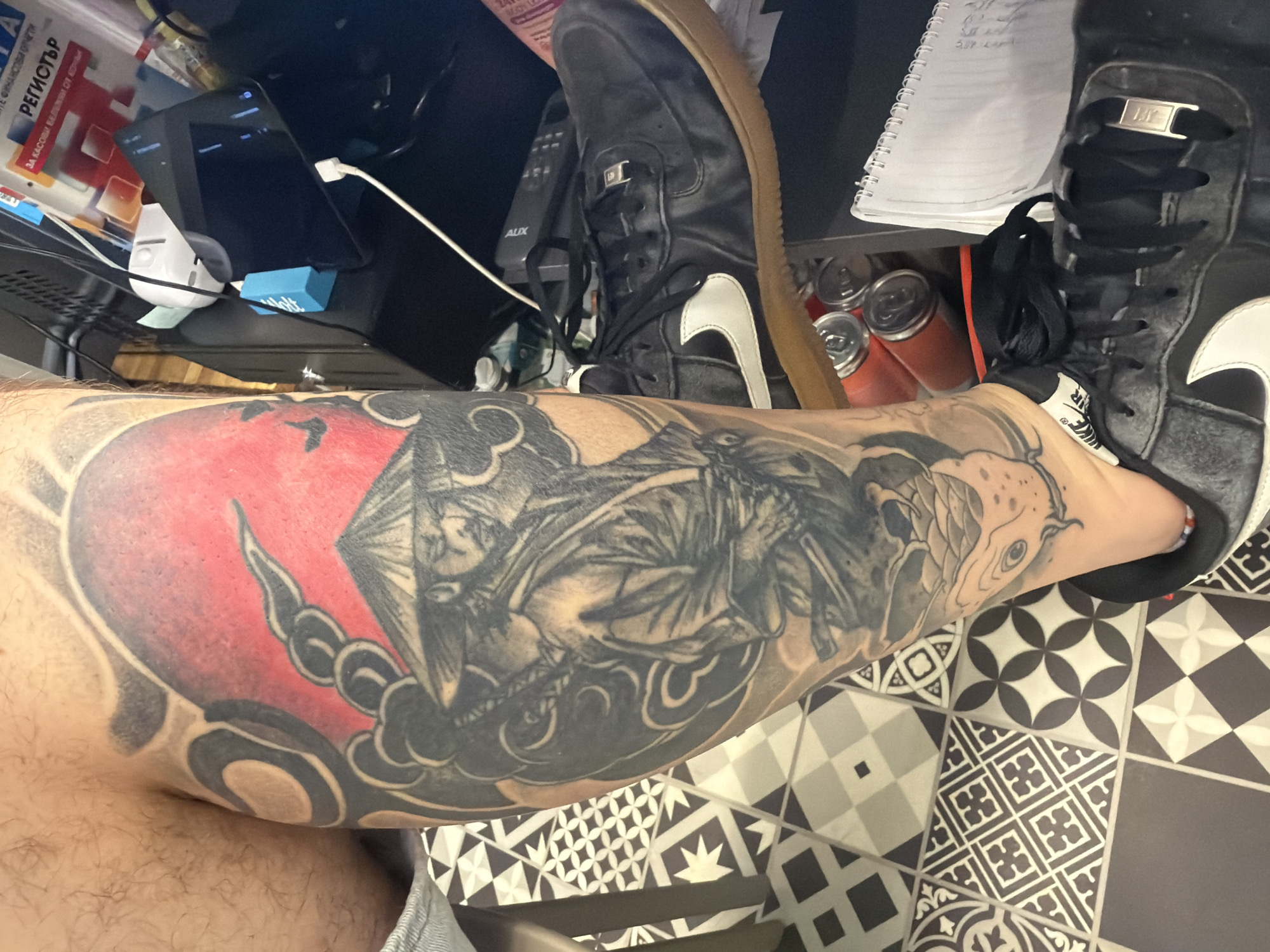 A partially visible adult male leg with a large Japanese-style tattoo depicting a samurai and other imagery, including a red sun, is in the foreground. The tattoo covers the majority of the lower leg, from thigh to ankle. The leg rests on a black and white patterned tile floor. Adjacent to the leg is a chair. A black shoe, possibly a Nike Air Force 1, is partially visible over the leg and tattoo. In the background, there is a cluttered desk area containing a variety of items, including cans, a notebook, a tablet, a computer, and assorted paperwork. The location appears to be indoors, likely a home or office. There is no explicit information to confirm the location as the city of Sofia, Bulgaria.