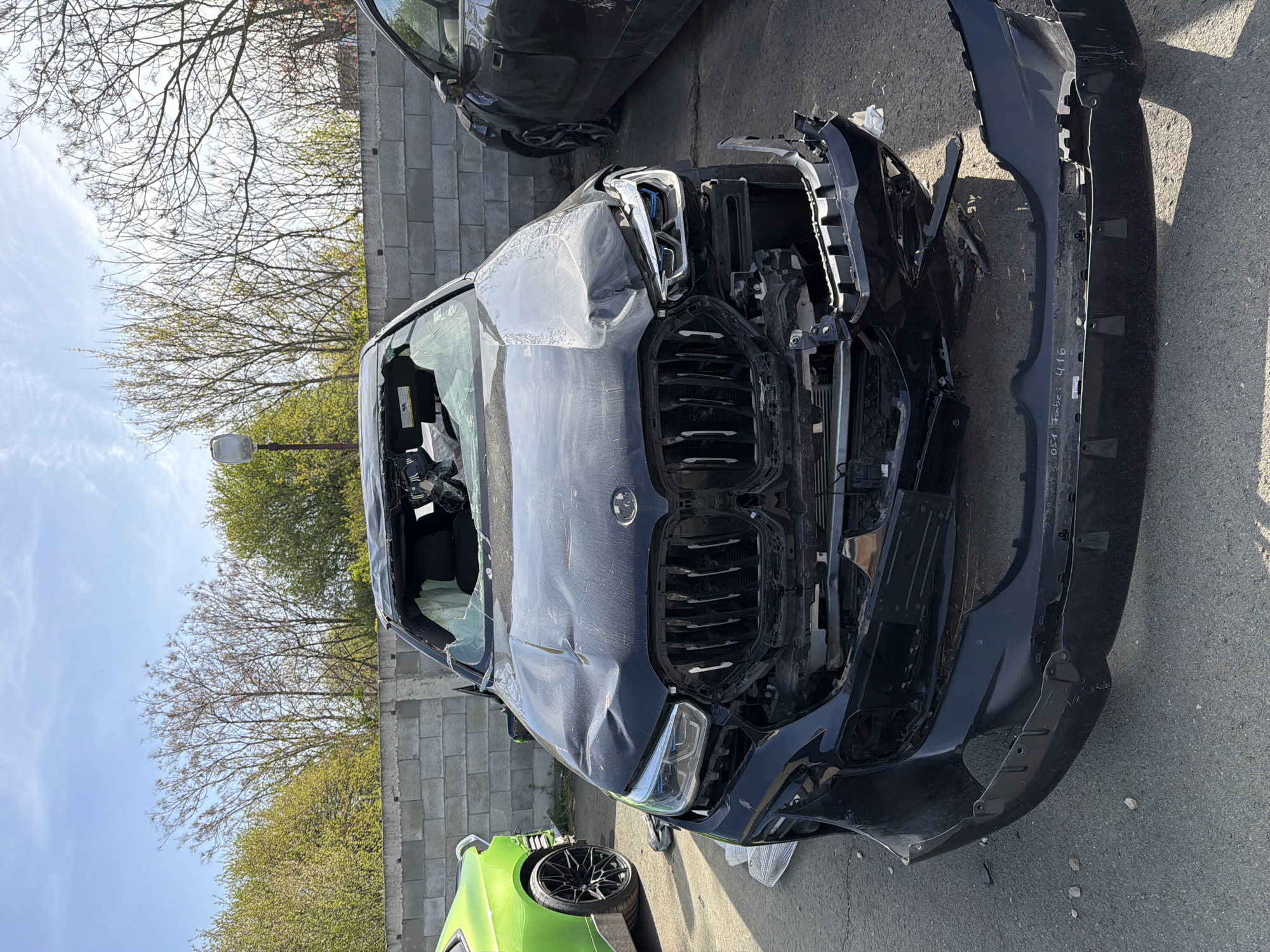 The image displays a heavily damaged black BMW vehicle, likely an SUV or SAV, in an outdoor setting that resembles a junkyard or vehicle storage area. The car has sustained severe frontal damage, with its hood significantly crumpled and deformed, exposing internal components. The front bumper is completely detached and lies on the ground next to the car, revealing the grille, which is also damaged and partially missing. The headlights appear to be broken or removed, and the radiator and other engine parts are visible.
The windshield on the driver's side is shattered or missing, and the driver's door window is also broken. Inside the vehicle, airbags appear to have deployed, indicated by white material visible within the cabin. The car's interior also shows disarray, suggesting the impact was significant.
In the background, another dark-colored car is partially visible. In the foreground, part of a bright green vehicle with a distinctive black multi-spoke wheel and tire is present. The ground is asphalt.
The setting includes a concrete block wall behind the damaged BMW. Above the wall, various trees are visible, some with bare branches and others with green foliage, indicating either late winter or early spring. A street light stands between the trees and the wall. The sky is bright blue with a few scattered clouds, suggesting a clear, sunny daytime. Shadows are visible on the ground, consistent with bright daylight.
On the detached black bumper, white handwritten text is partially legible, including "054 Fabc 416". A warning label on the passenger sun visor inside the damaged car is visible but unreadable. No direct activity is occurring in the scene; it depicts the aftermath of a severe vehicular accident.